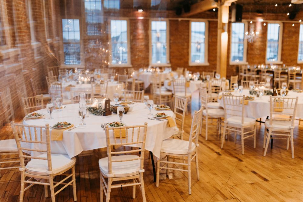 Bright rustic wedding reception room with round white-draped tables, chiavari chairs, and exposed brick.