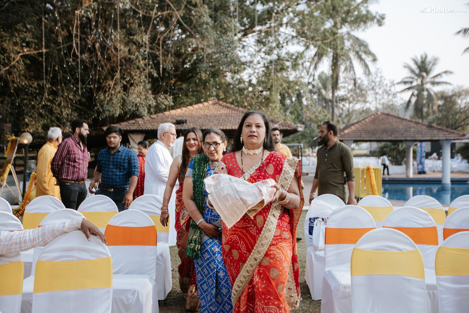 Outdoor Indian wedding celebration with colorfully dressed guests and decorated white chairs.