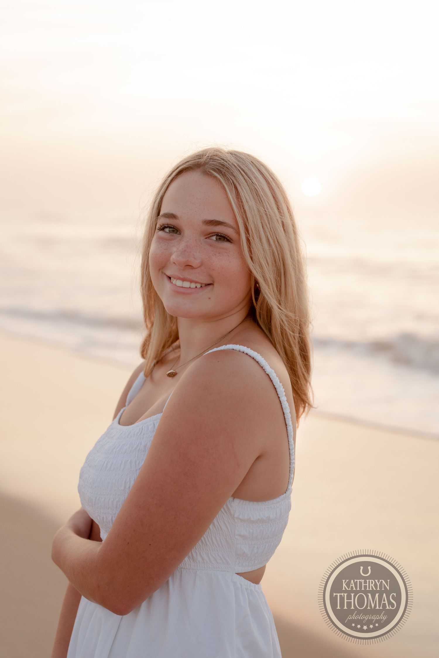 A person in a white dress poses for a beach portrait at sunset with a warm glowing sky in the background.