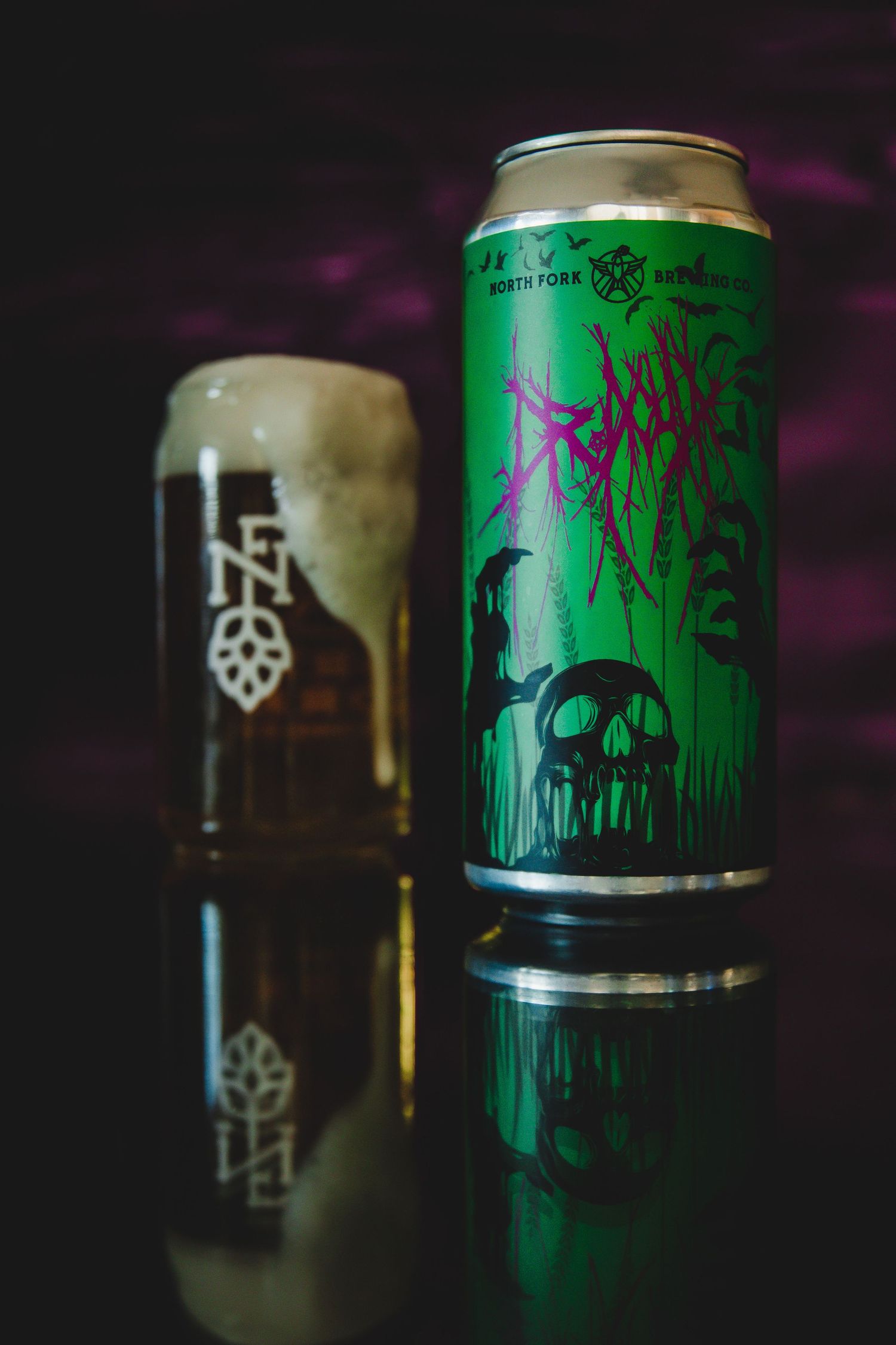 Green horror-themed craft beer can with skull artwork next to a pint glass on reflective surface.
