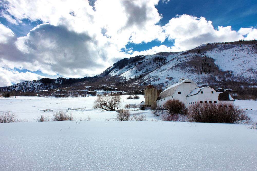 Snow covered winter landscape with mountains and scattered buildings under dramatic cloudy sky.
