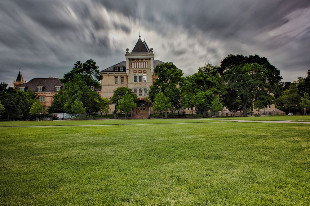 Grand historic building with tower on green lawn under dramatic cloudy sky.