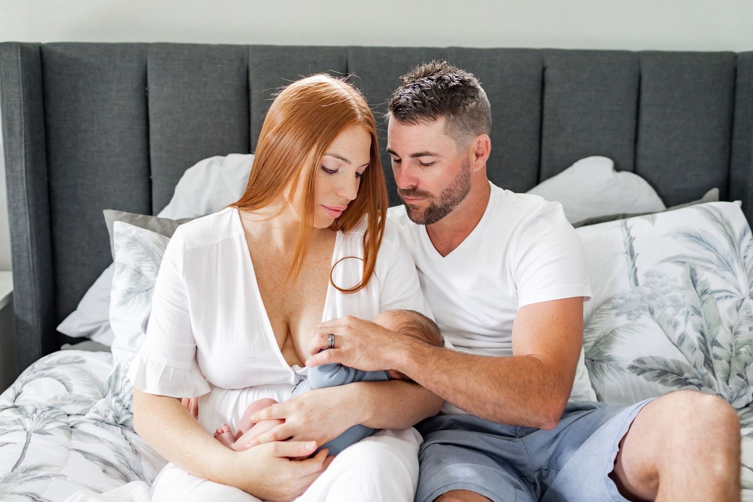 Parents sitting on gray bed gently hold their newborn baby.