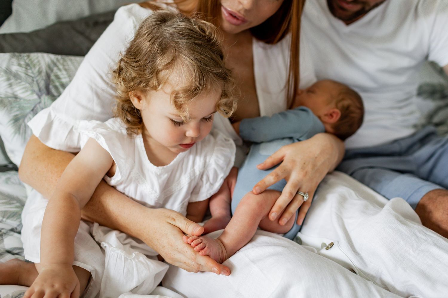 Family bonding moment on bed as child touches newborn sibling's feet.