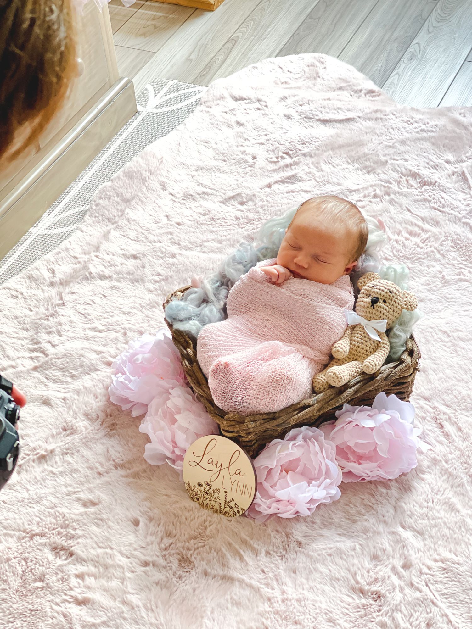 Sleeping newborn wrapped in pink swaddle lying in basket with floral decorations and teddy bear.