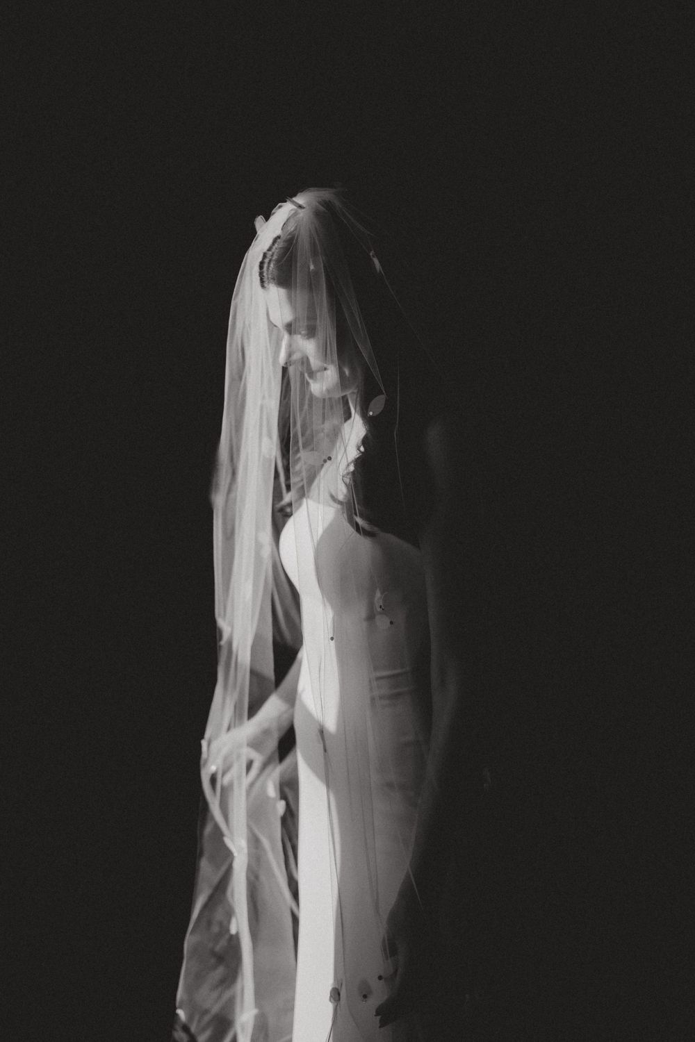 Elegant bride in fitted white dress and flowing veil poses in dramatic black and white portrait.