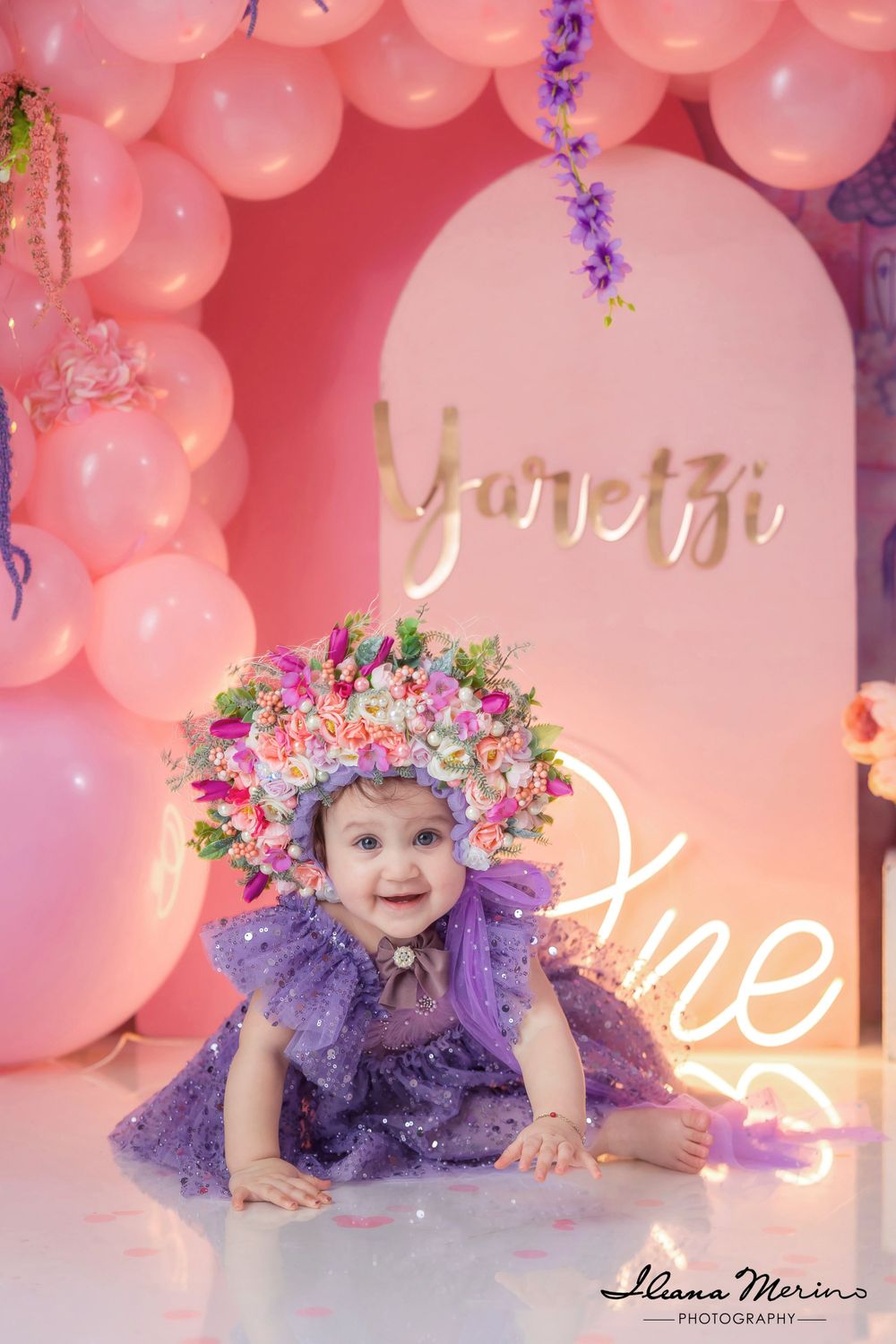 Baby girl in purple dress with floral crown smiles at first birthday party celebration.