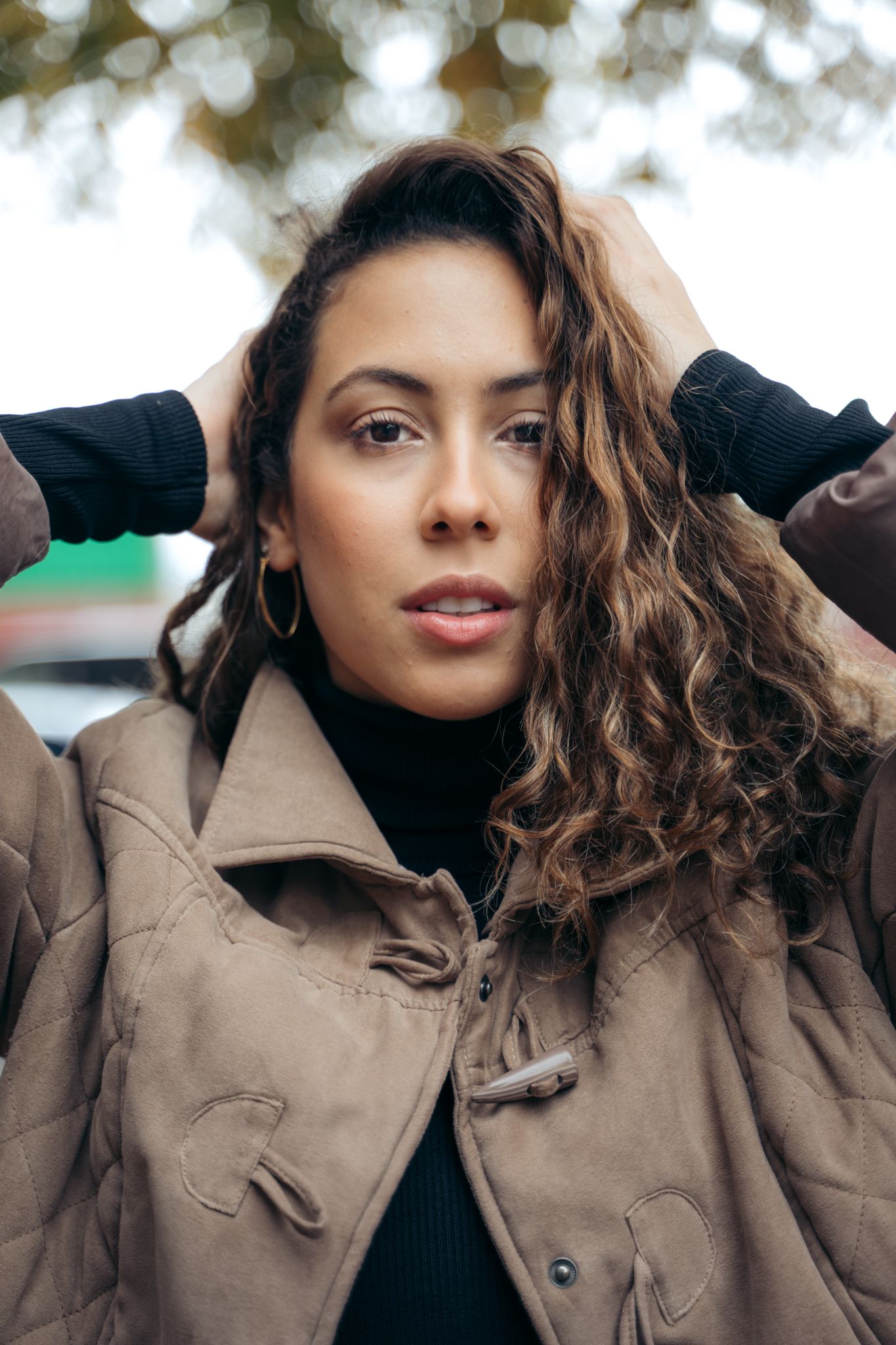 Young woman with curly hair posing outdoors in beige jacket and black turtleneck.