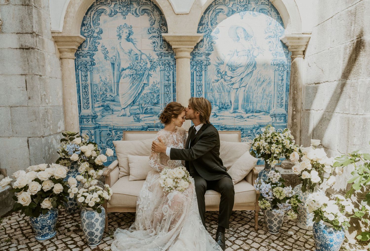 Romantic wedding couple seated against blue and white Portuguese azulejo tiles surrounded by white flowers.