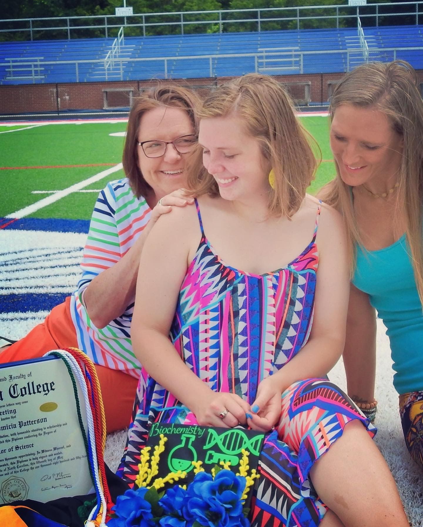 Three people smiling together on a blue sports field with graduation materials.