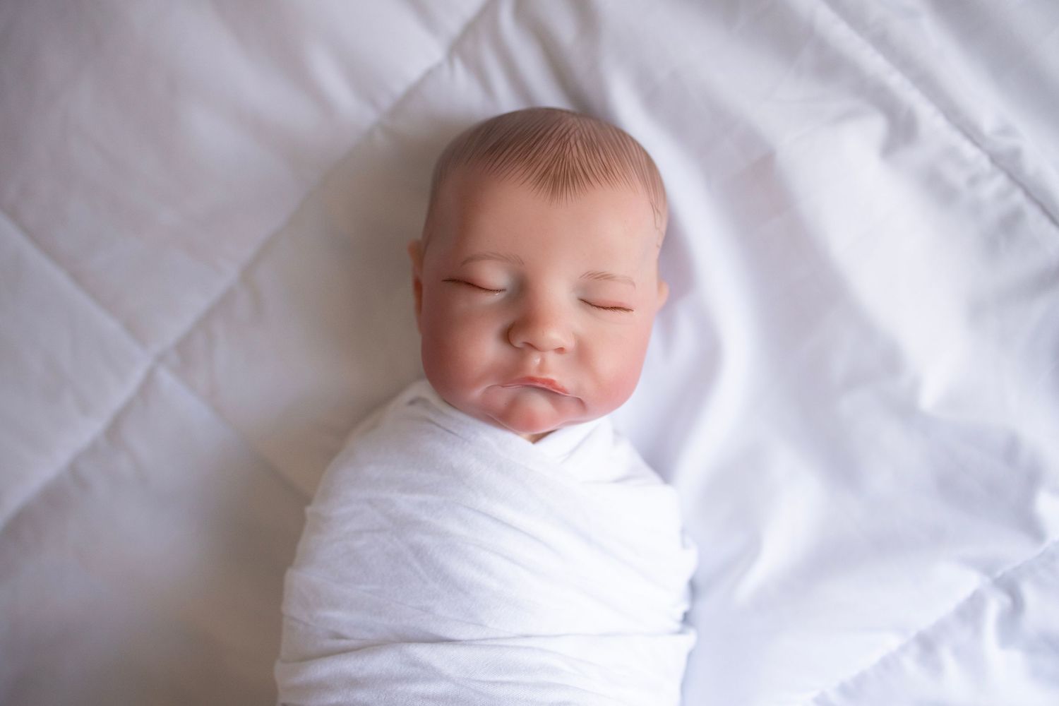 Baby doll swaddled in a white wrap laying on a simple white bed cover.
