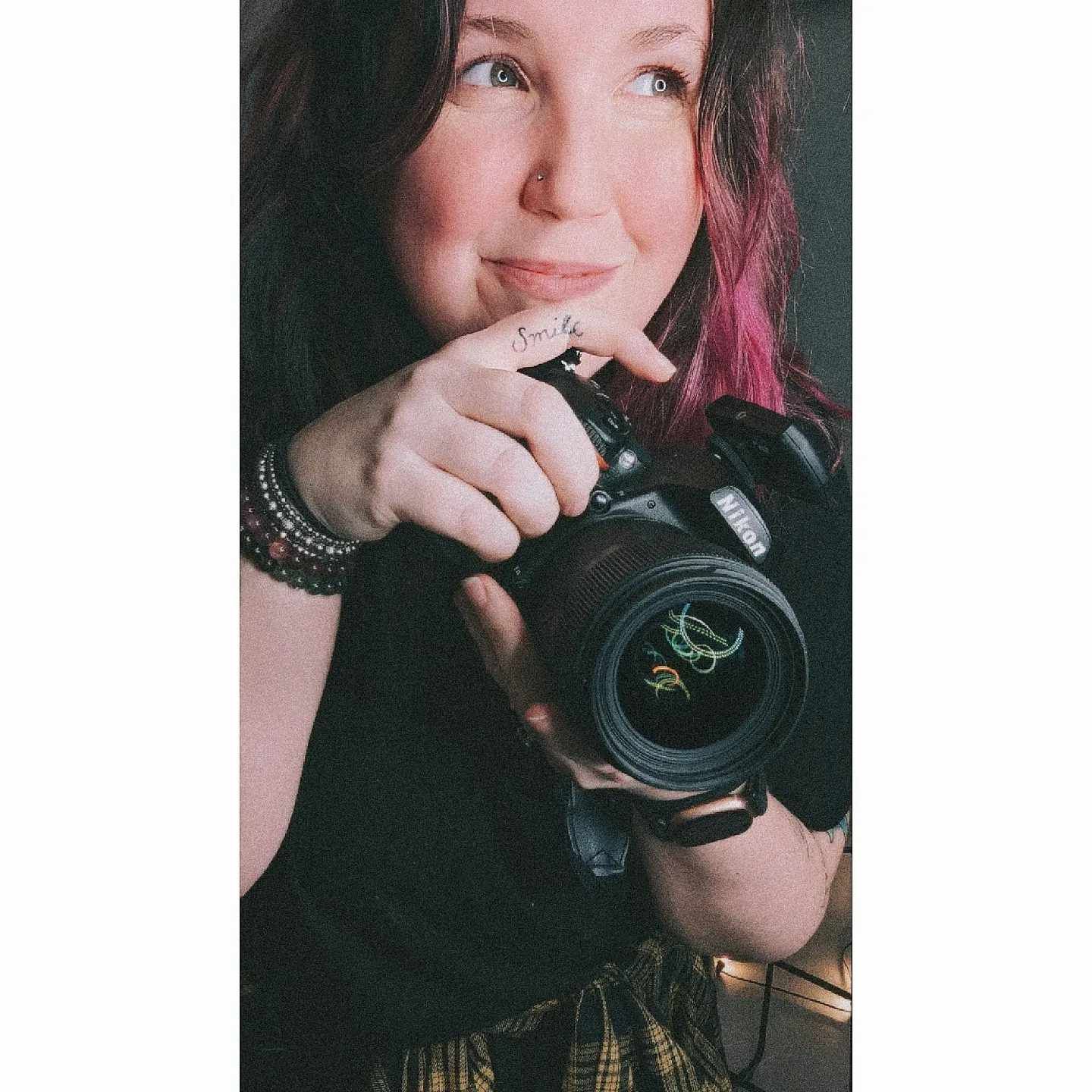 Female photographer smiling and holding DSLR camera with pink highlighted hair and layered bracelets.