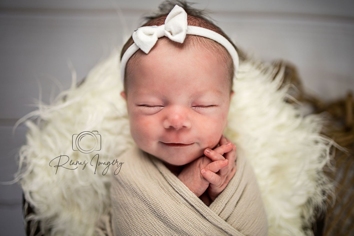 Sleeping newborn baby girl in cream wrap with white bow headband on fluffy blanket.