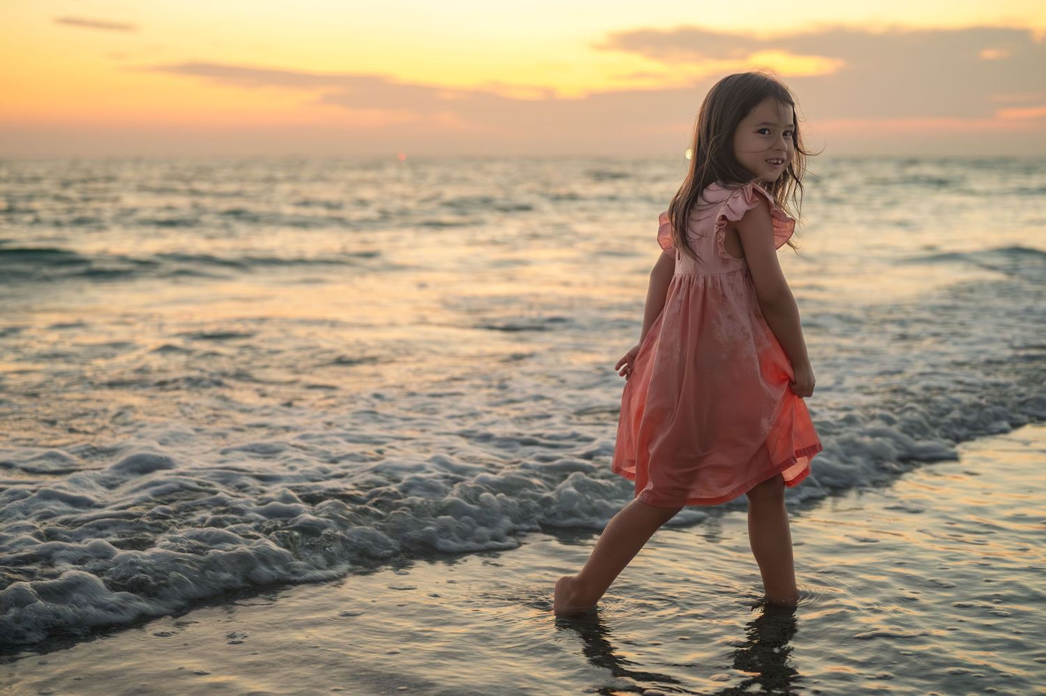 A child in a pink dress dancing at the beach during golden sunset.