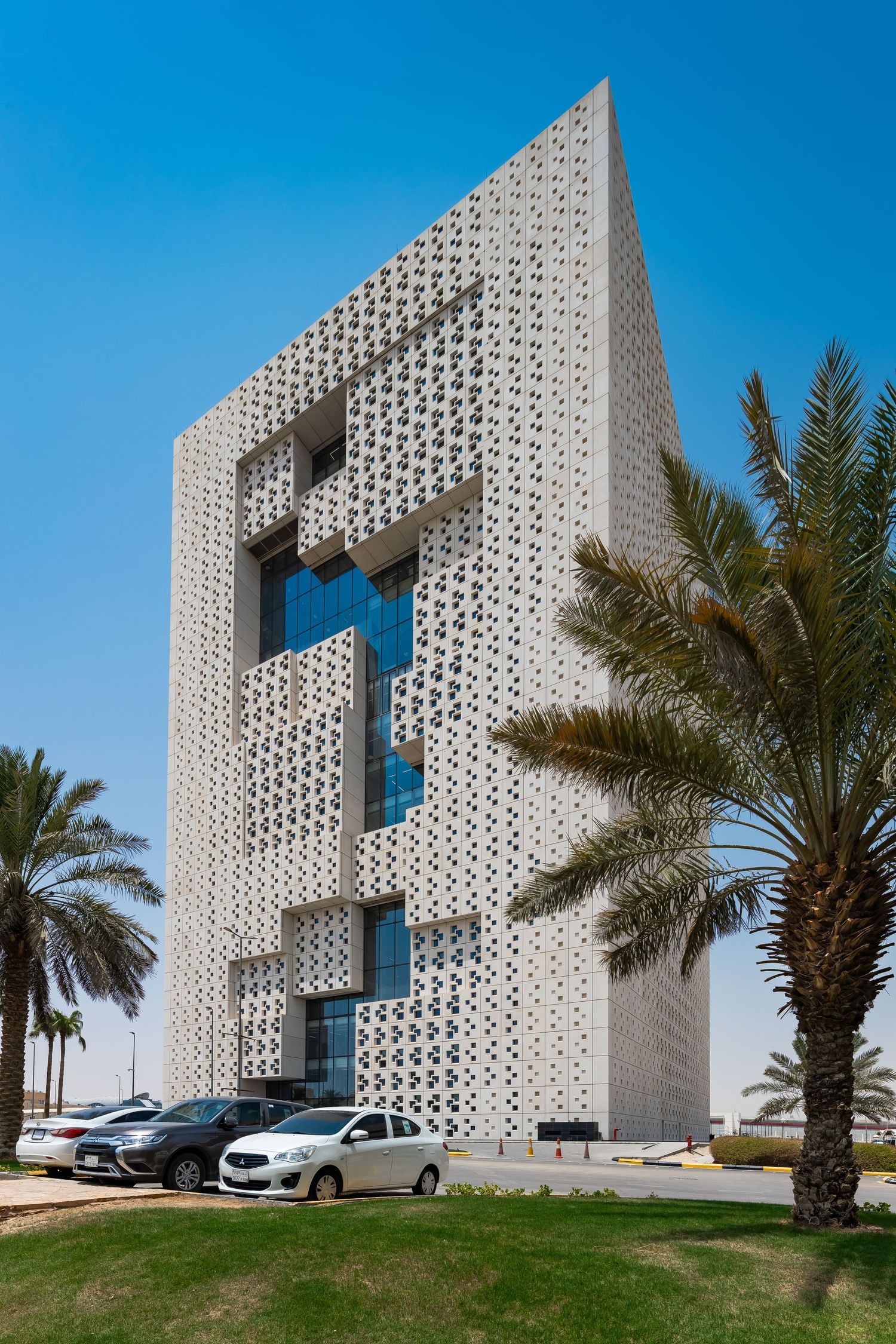 Modernist tower with distinctive lattice facade surrounded by palm trees and parked cars.