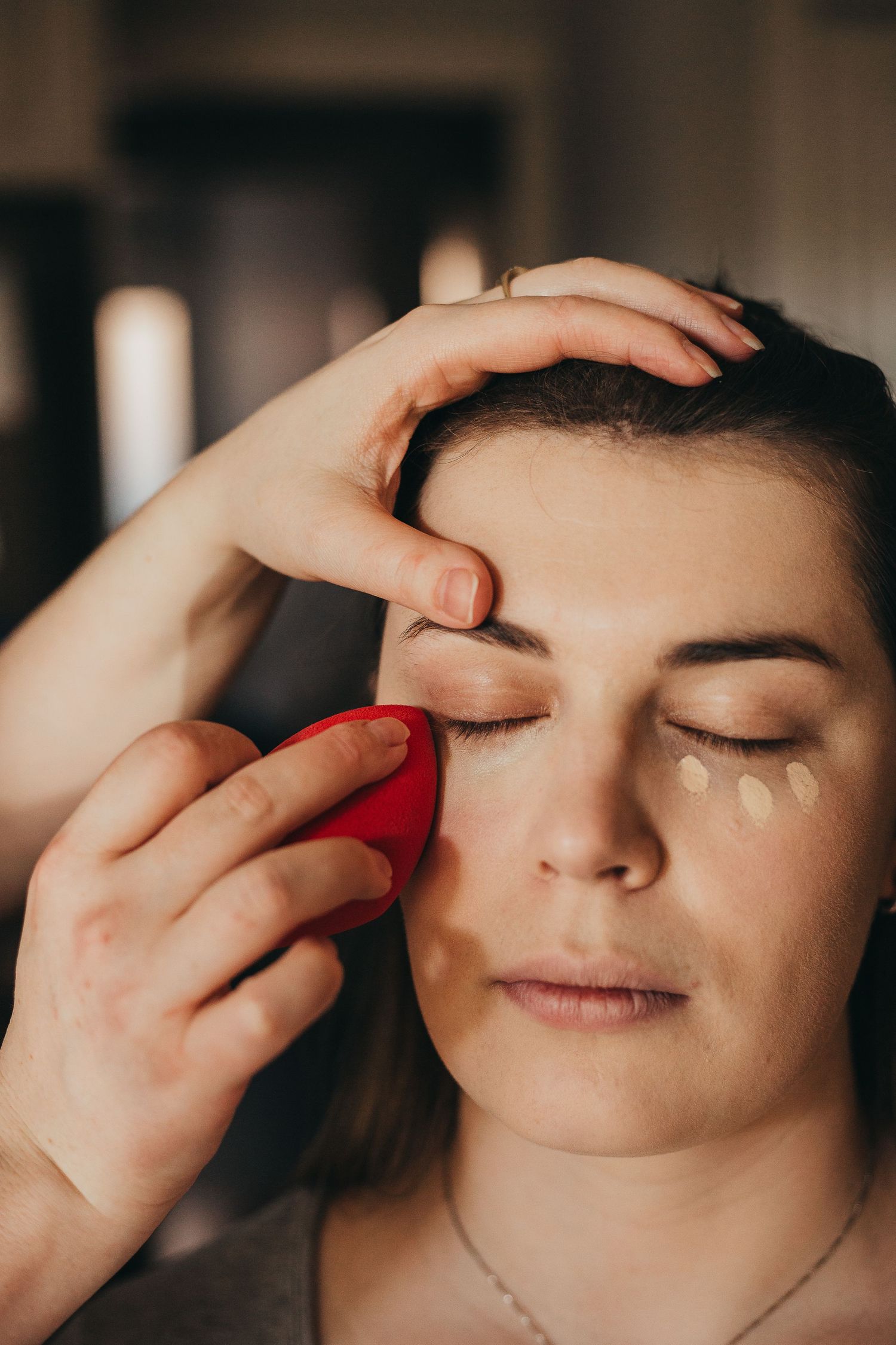 Close-up of makeup application using a red beauty blender sponge with concealer dots under eye.