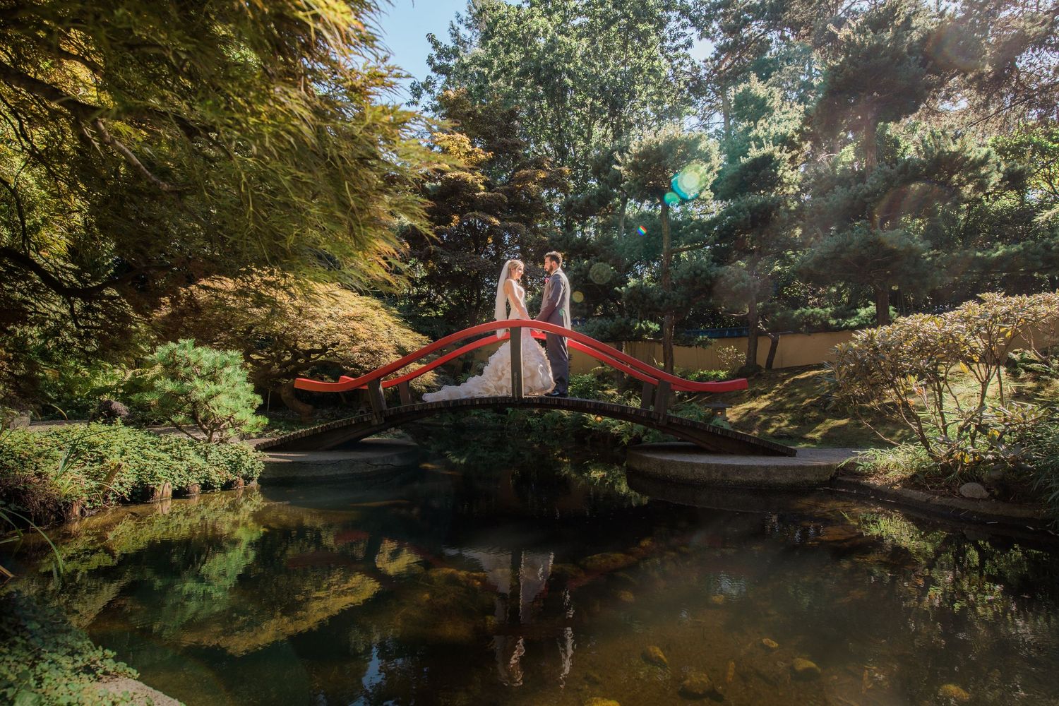 A romantic portrait on a red bridge over a reflective pond in a garden setting.