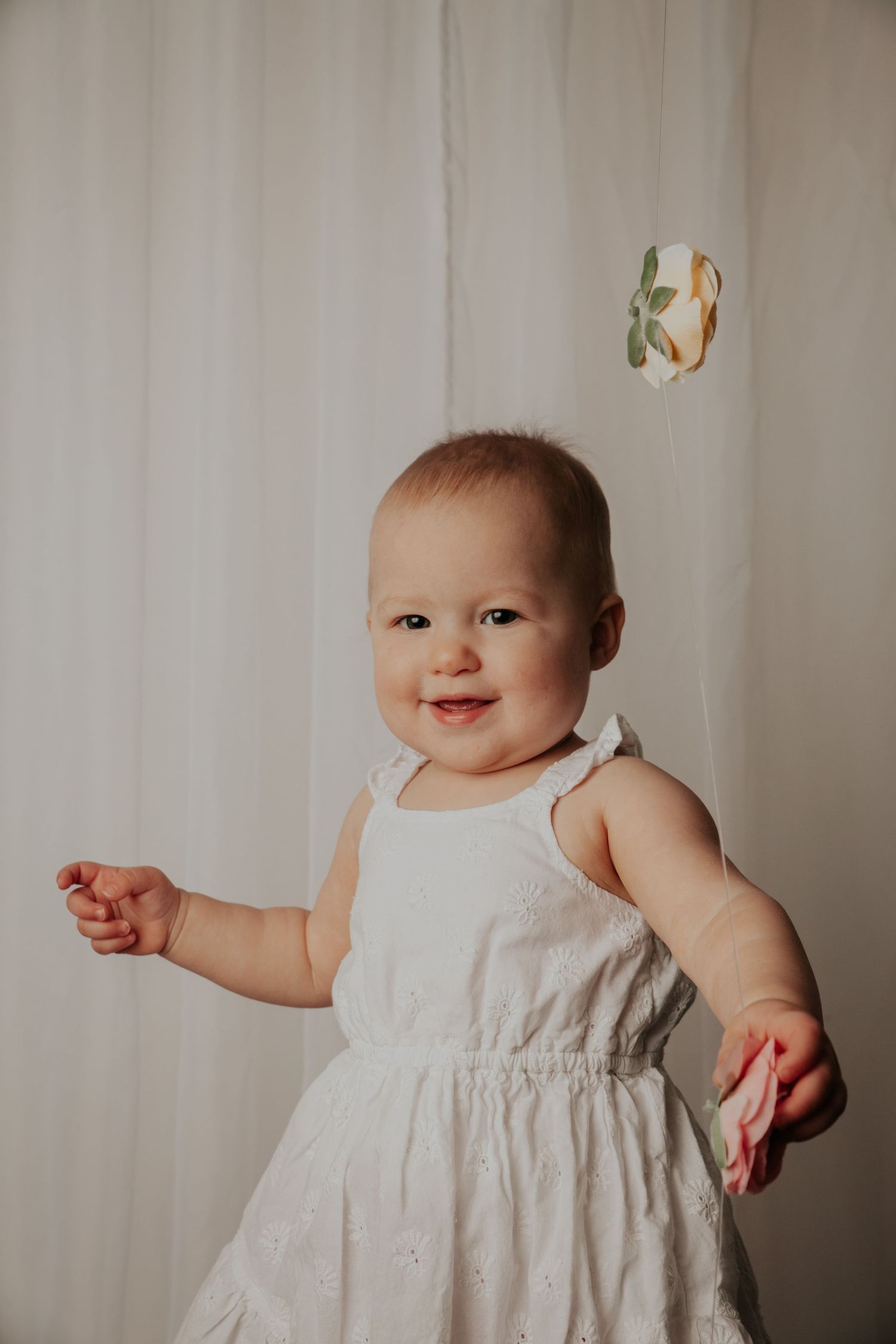 Smiling baby in white dress poses against light curtain background.