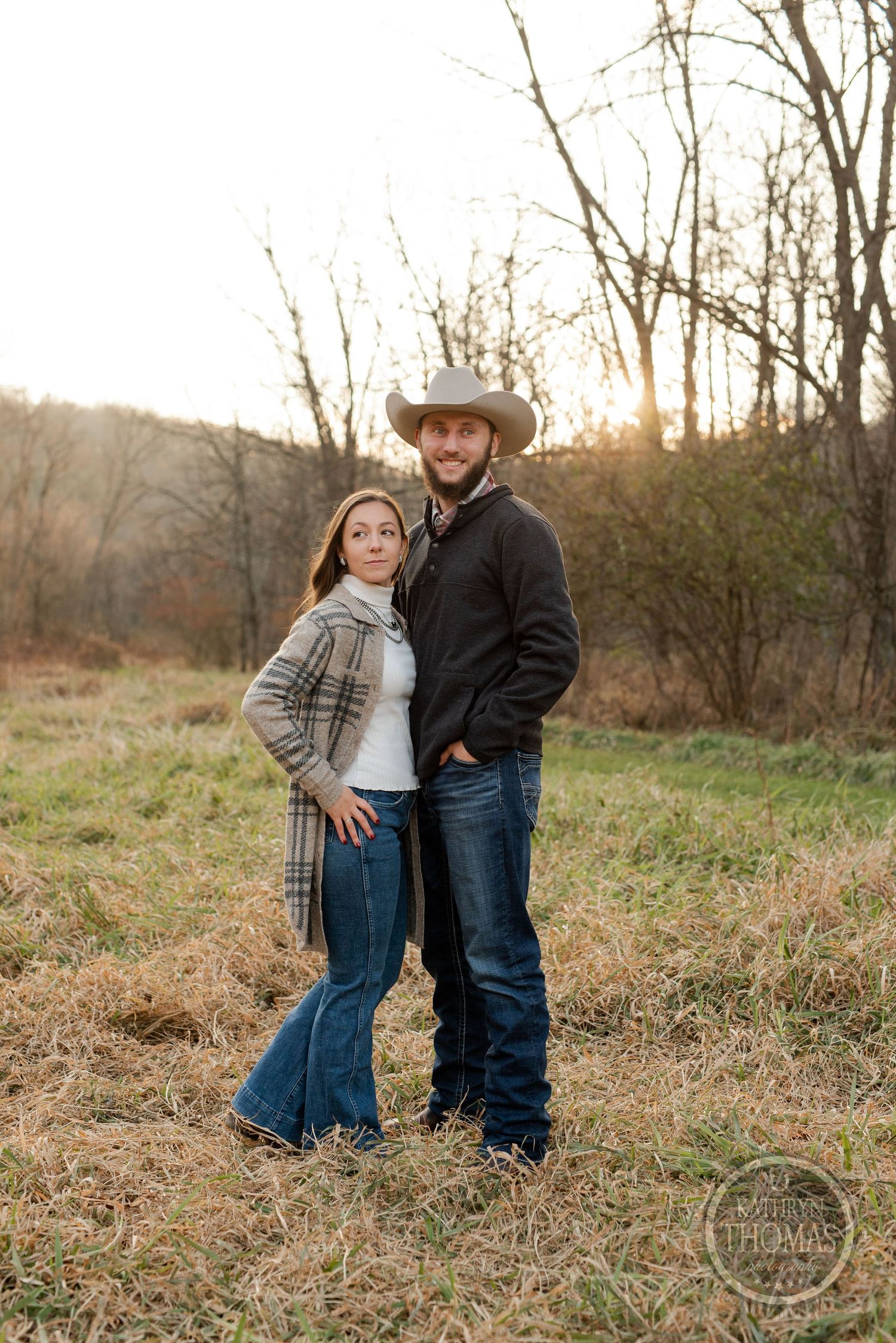 A couple in western attire poses together in a rustic field at sunset.