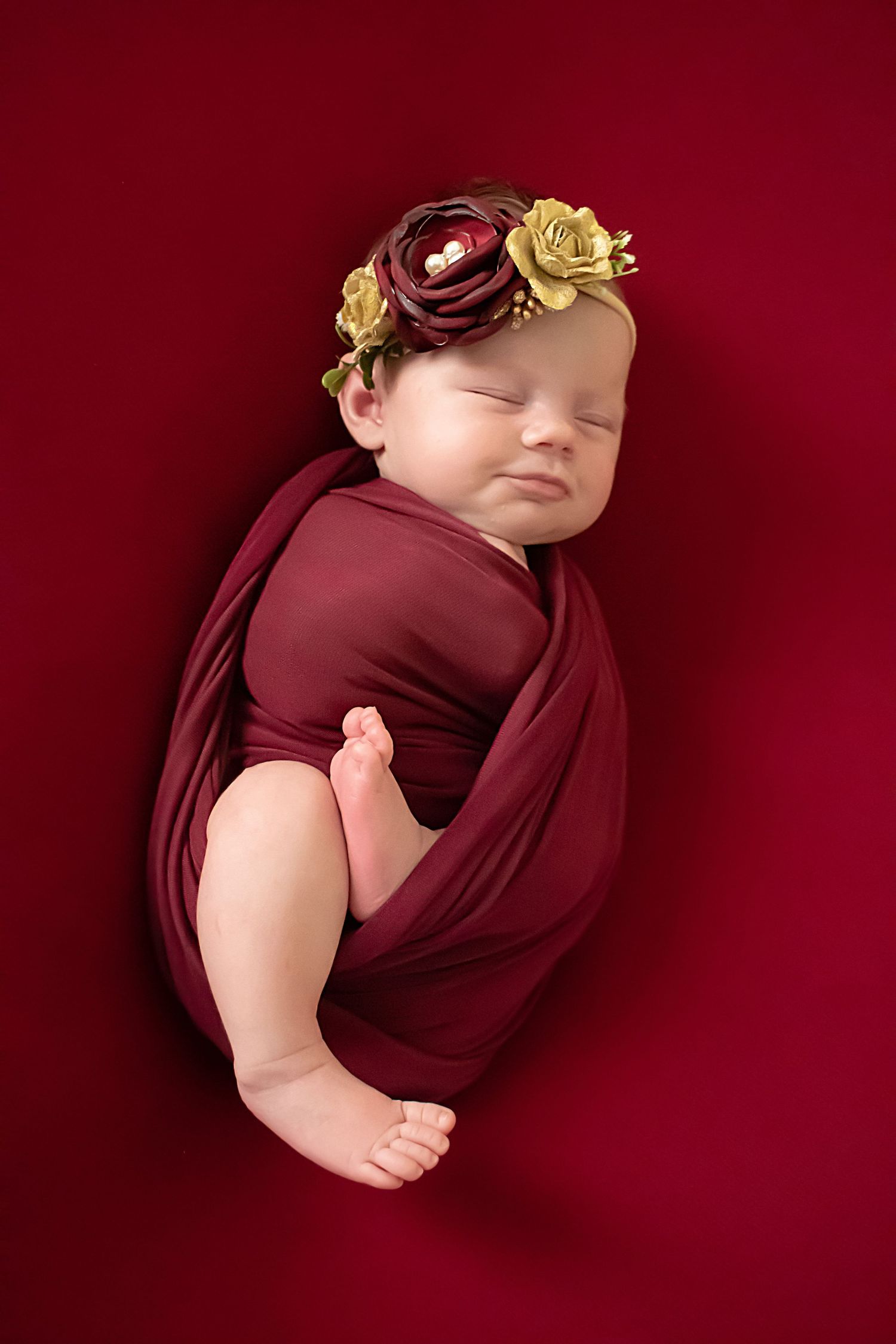 Sleeping newborn baby wrapped in burgundy fabric wearing floral headband with burgundy and gold flowers on red background.