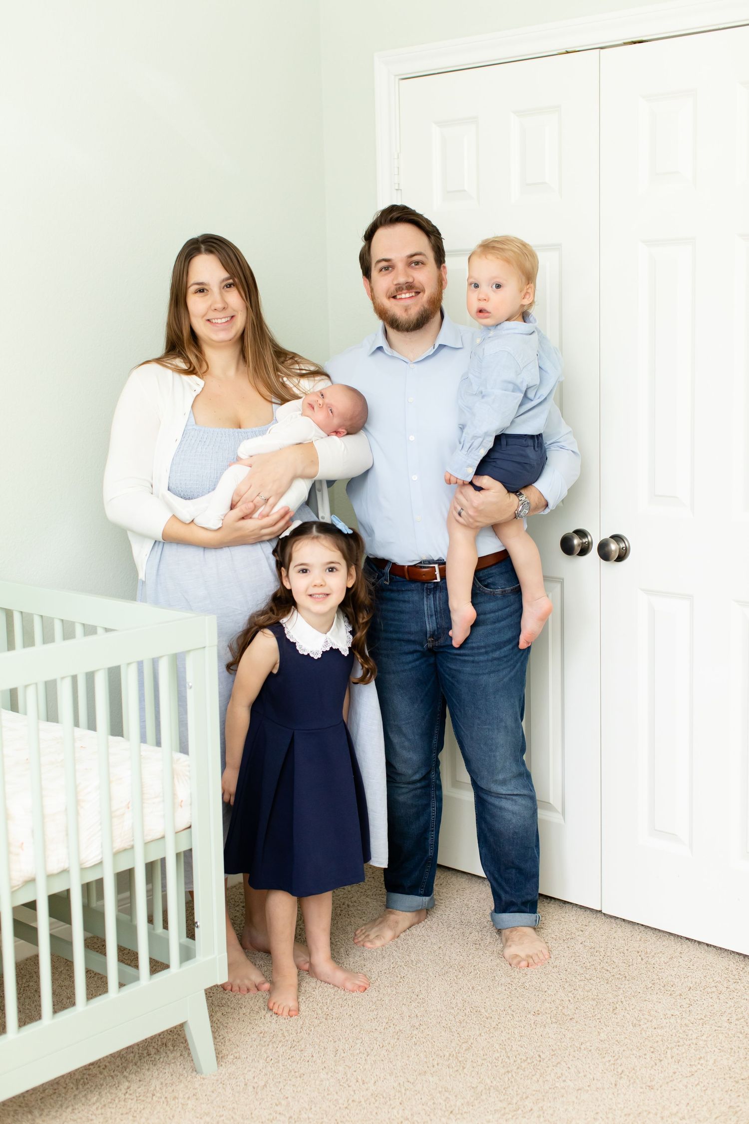 A growing family poses together in a nursery with mint green walls and white furniture.