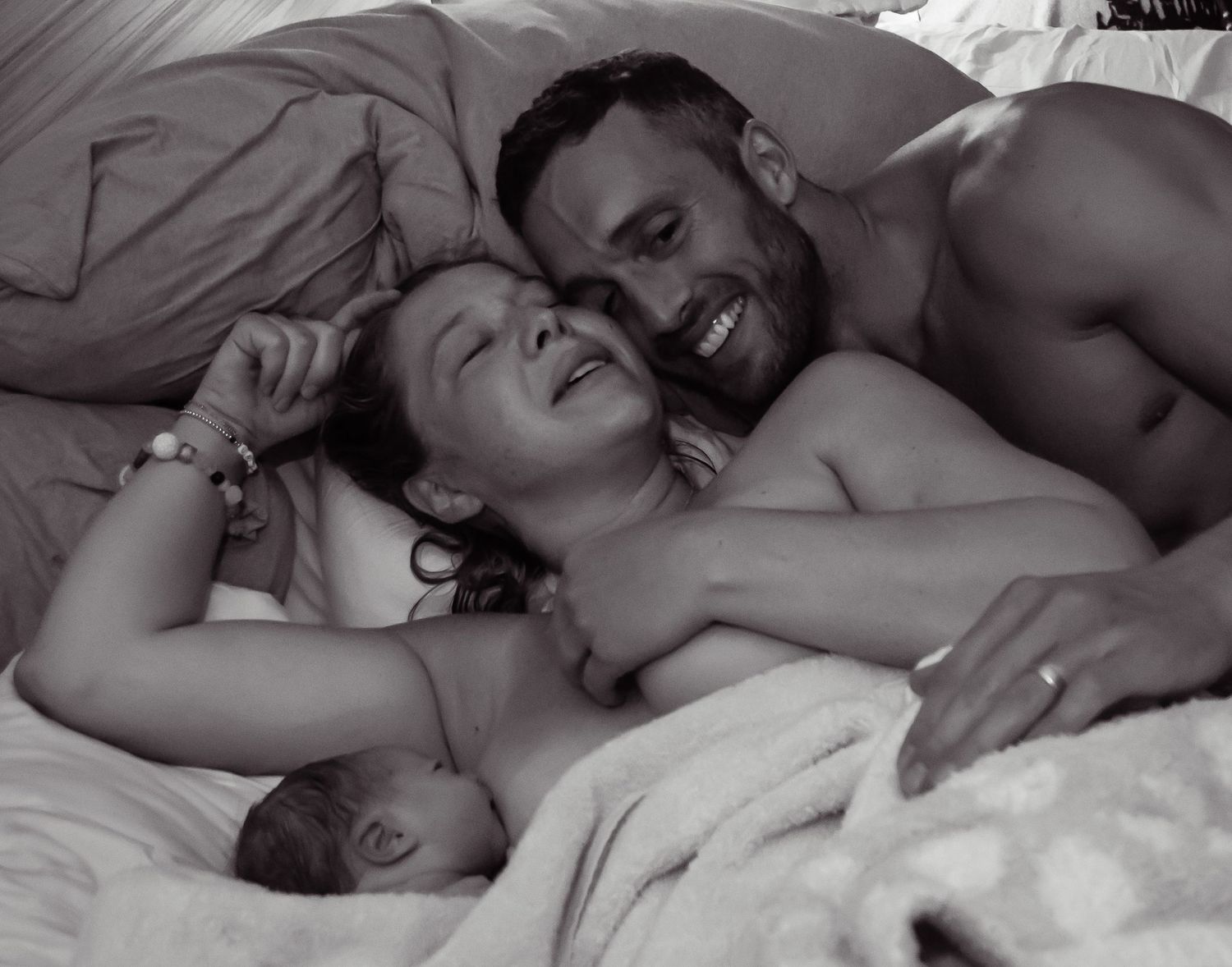 Image of a father cuddling his wife in a bed as she breastfeeds their newborn baby.