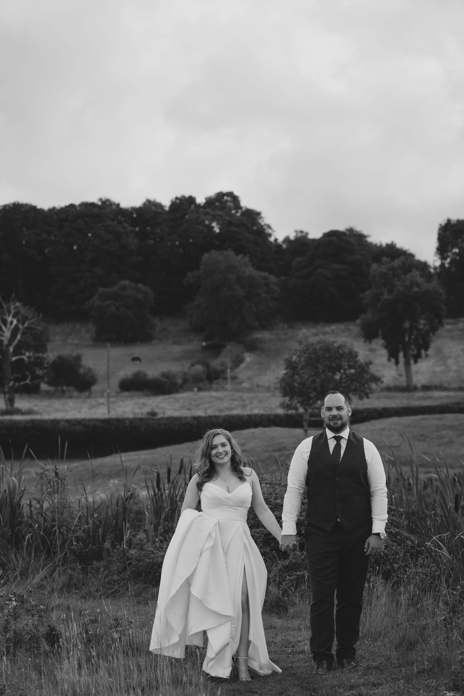 A black and white wedding portrait of a couple walking hand in hand through a rolling landscape.