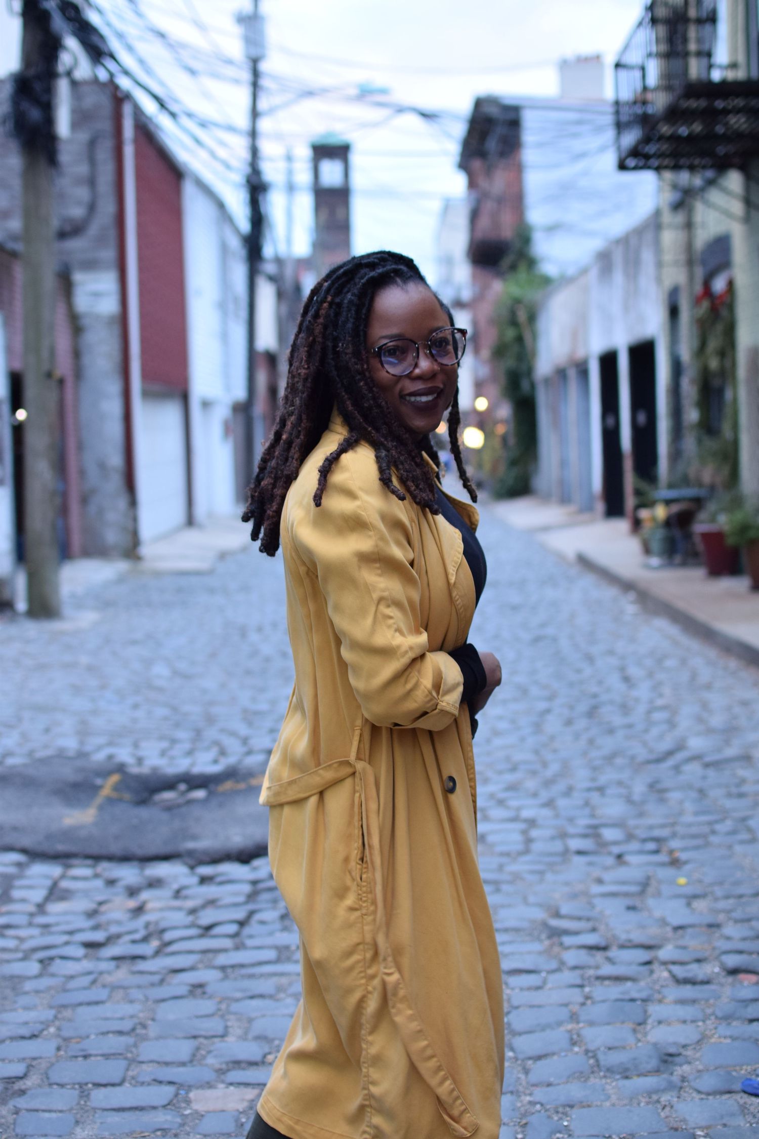 Person in a stylish yellow coat poses on a cobblestone street with historic buildings in the background.