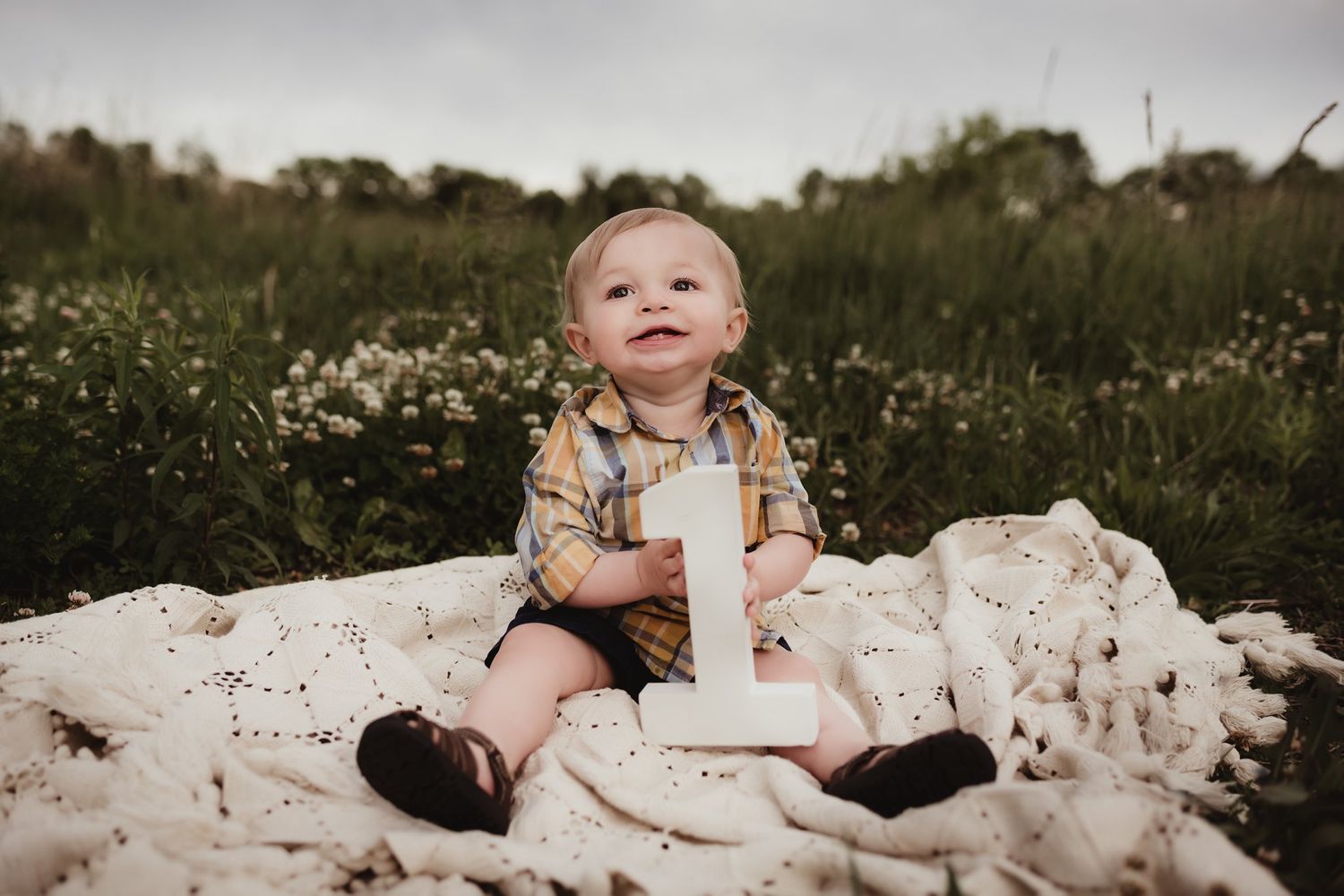 Baby wearing plaid shirt sits on white blanket in field of wildflowers with number one prop.