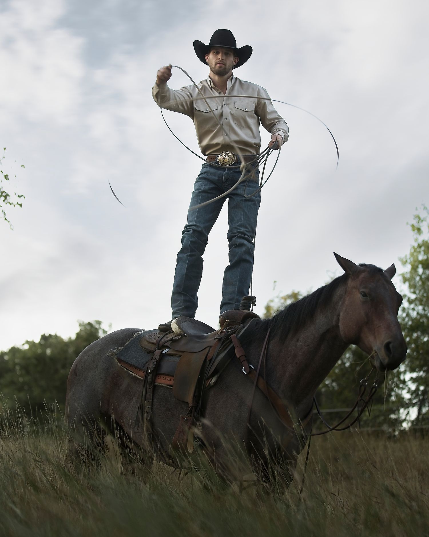 A rider stands on horseback while practicing with a lasso against a cloudy sky.