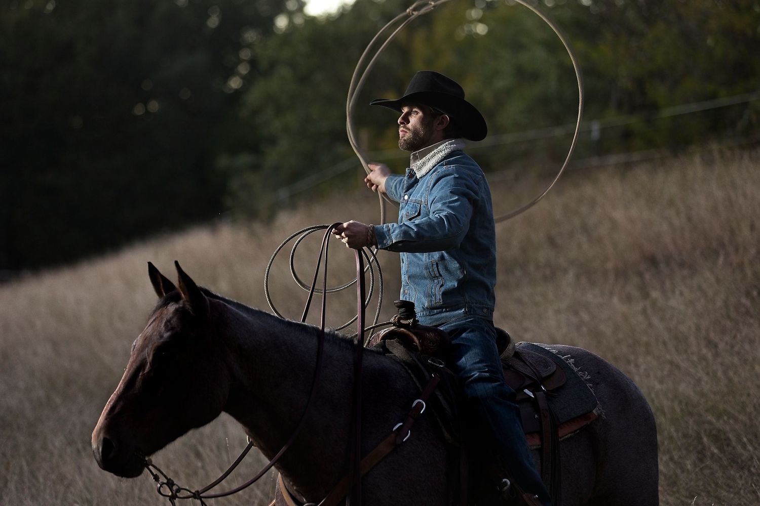 A rider in denim swings a lasso while mounted on horseback at dusk.