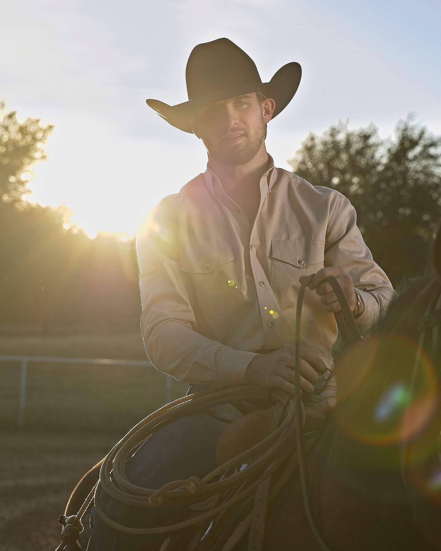 A cowboy in Western attire holds a lasso while backlit by golden sunset light.