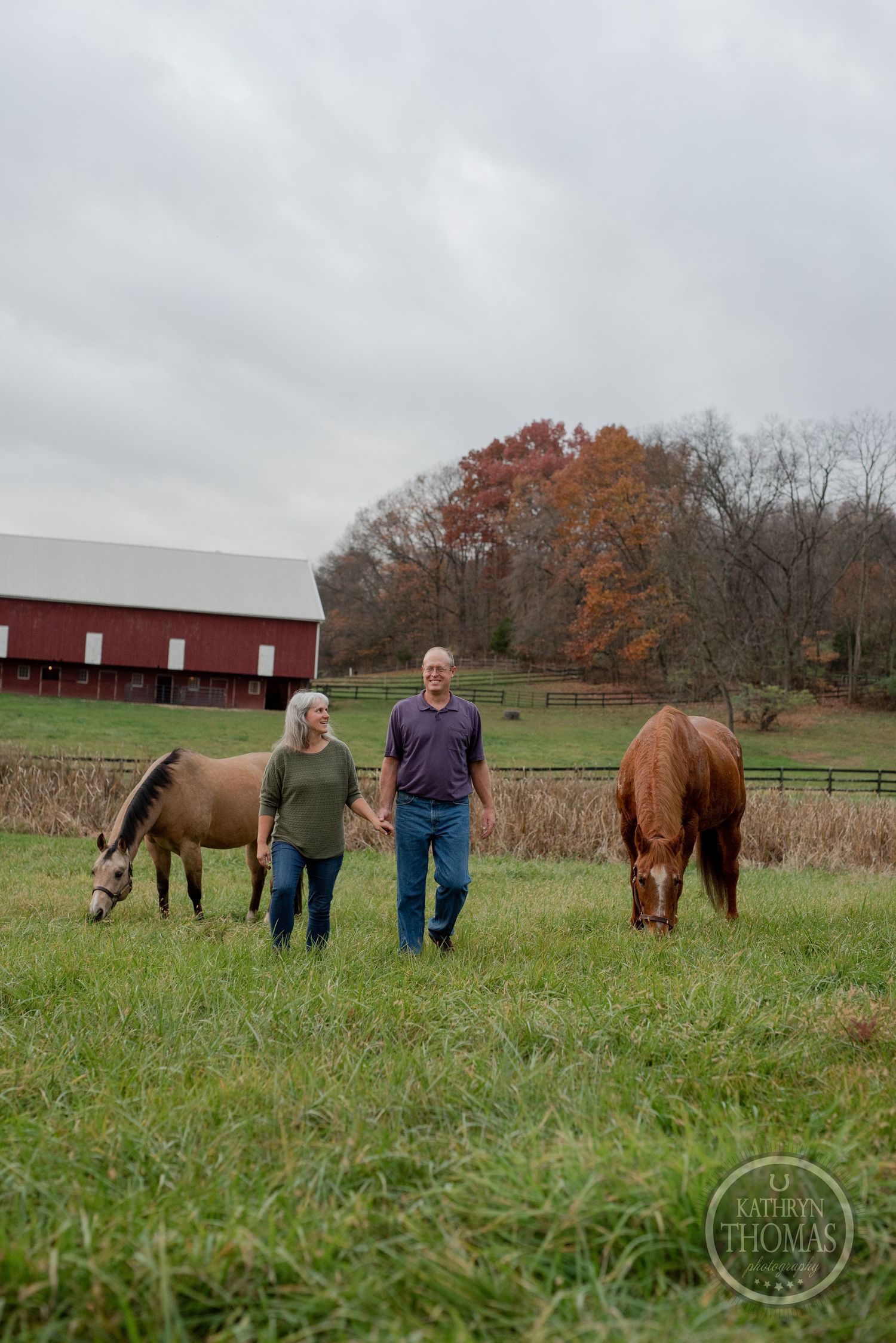 Two people stand between grazing horses in a pasture with red barn and fall foliage.