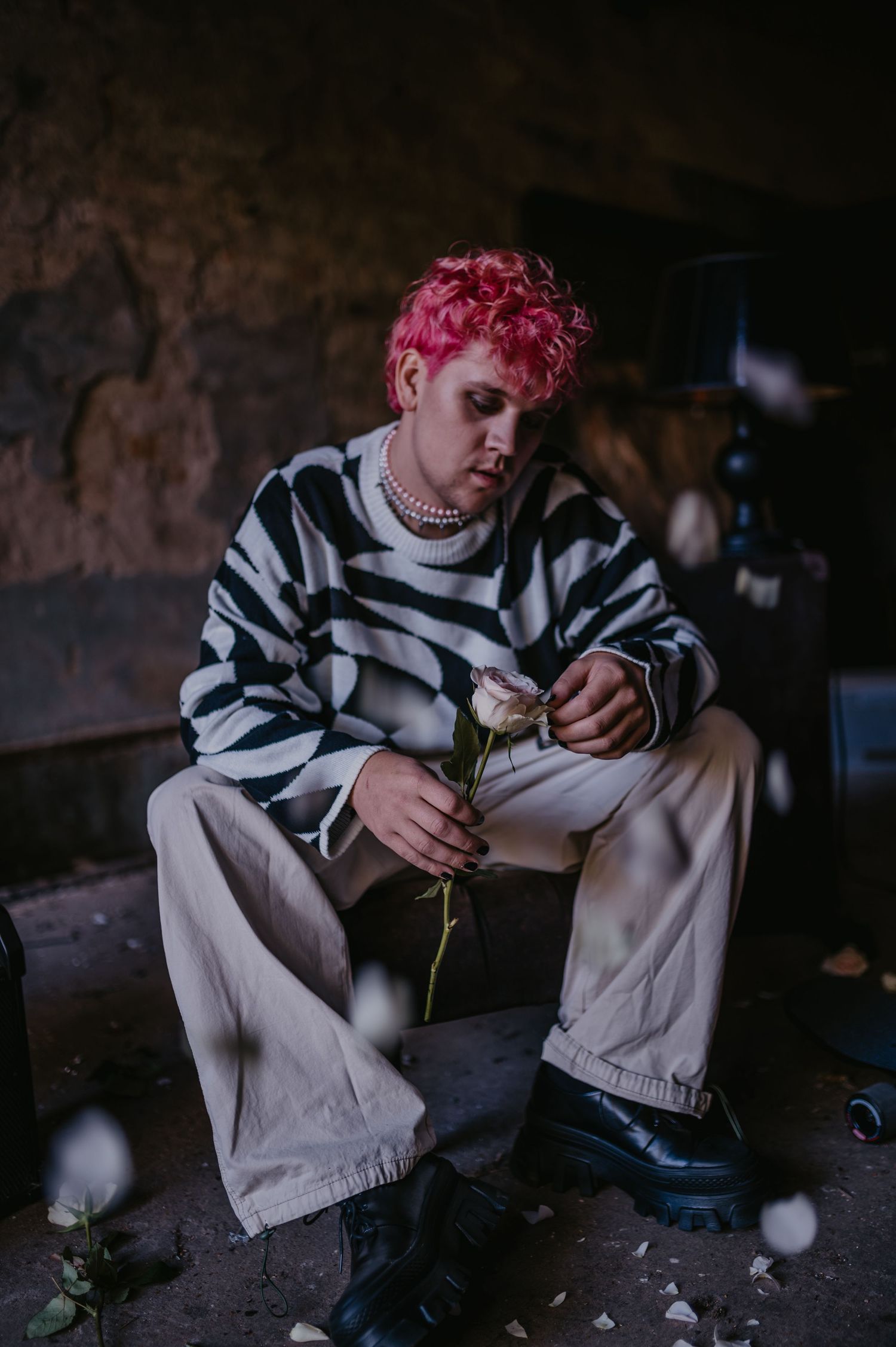 Person with pink curly hair in striped sweater holding a white rose while sitting in a dark abandoned space.