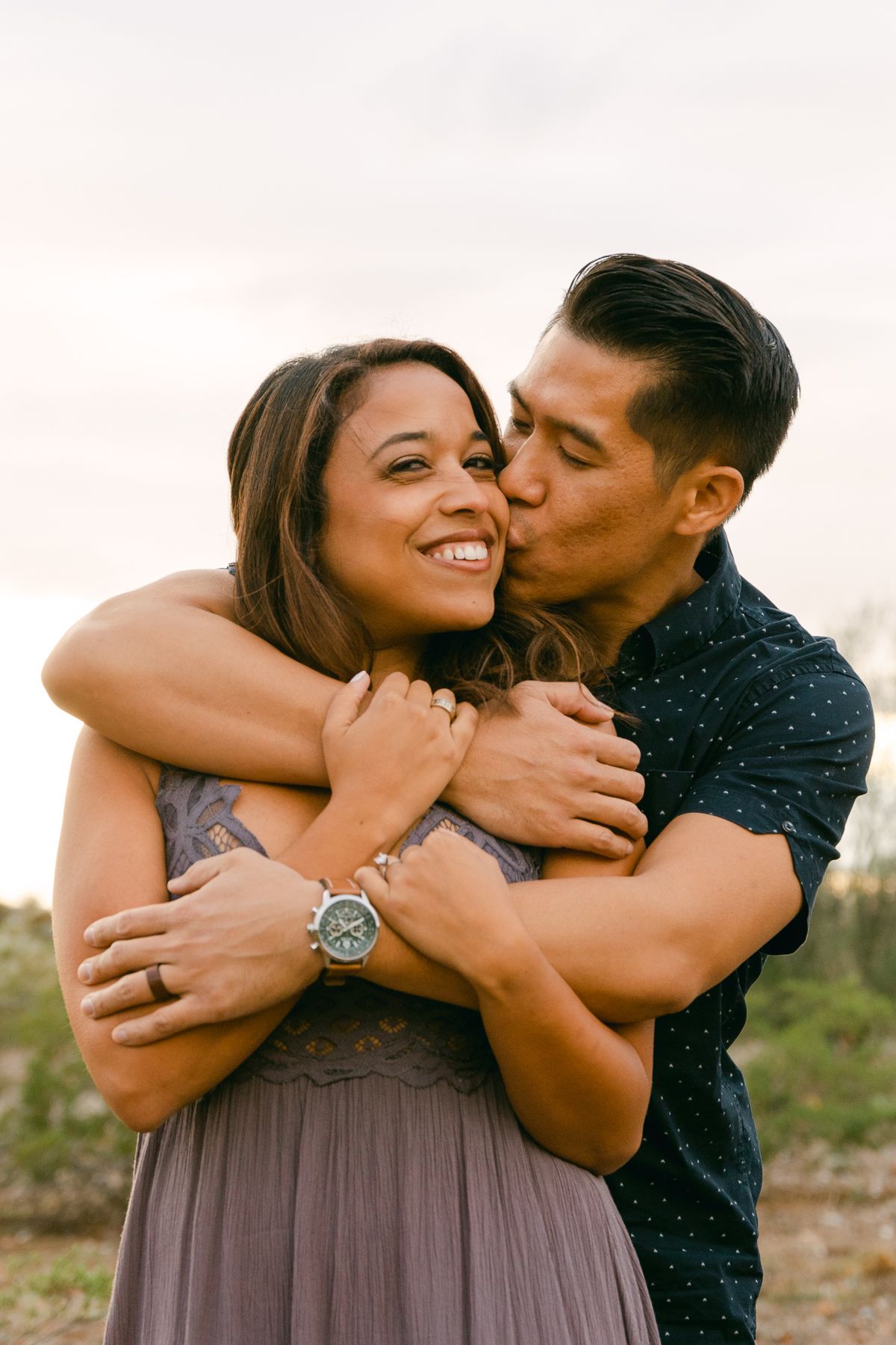 A man in black has his arms around a woman and he's kissing her cheek. She's smiling and there's greenery and dirt behind them.