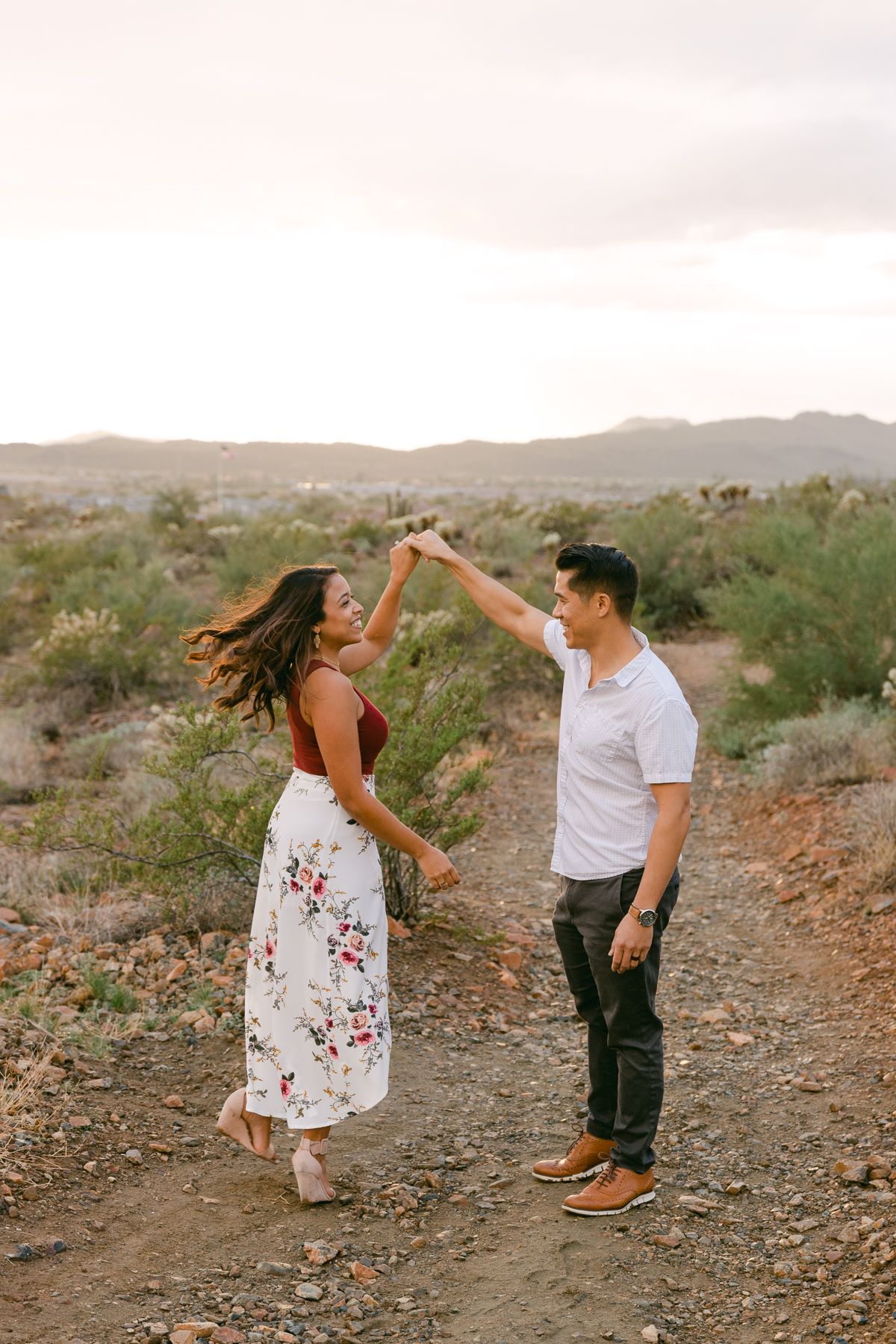 A man is twirling a woman on a desert path. Her skirt and hair is flowing and they're smiling. There's desert and mountains behind them.