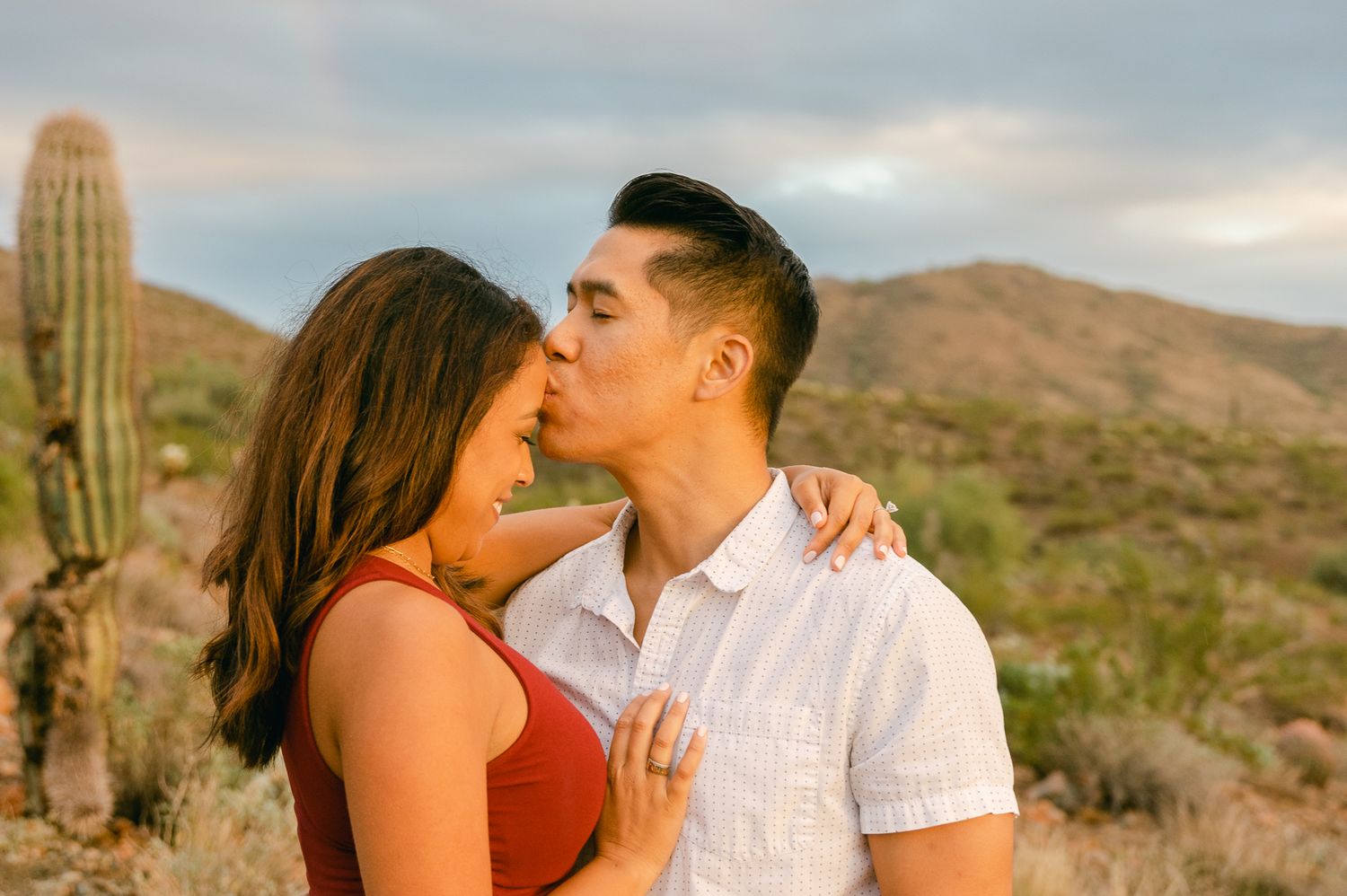 A man is kissing the forehead of a woman. She's smiling and there's a cactus and mountains behind them.