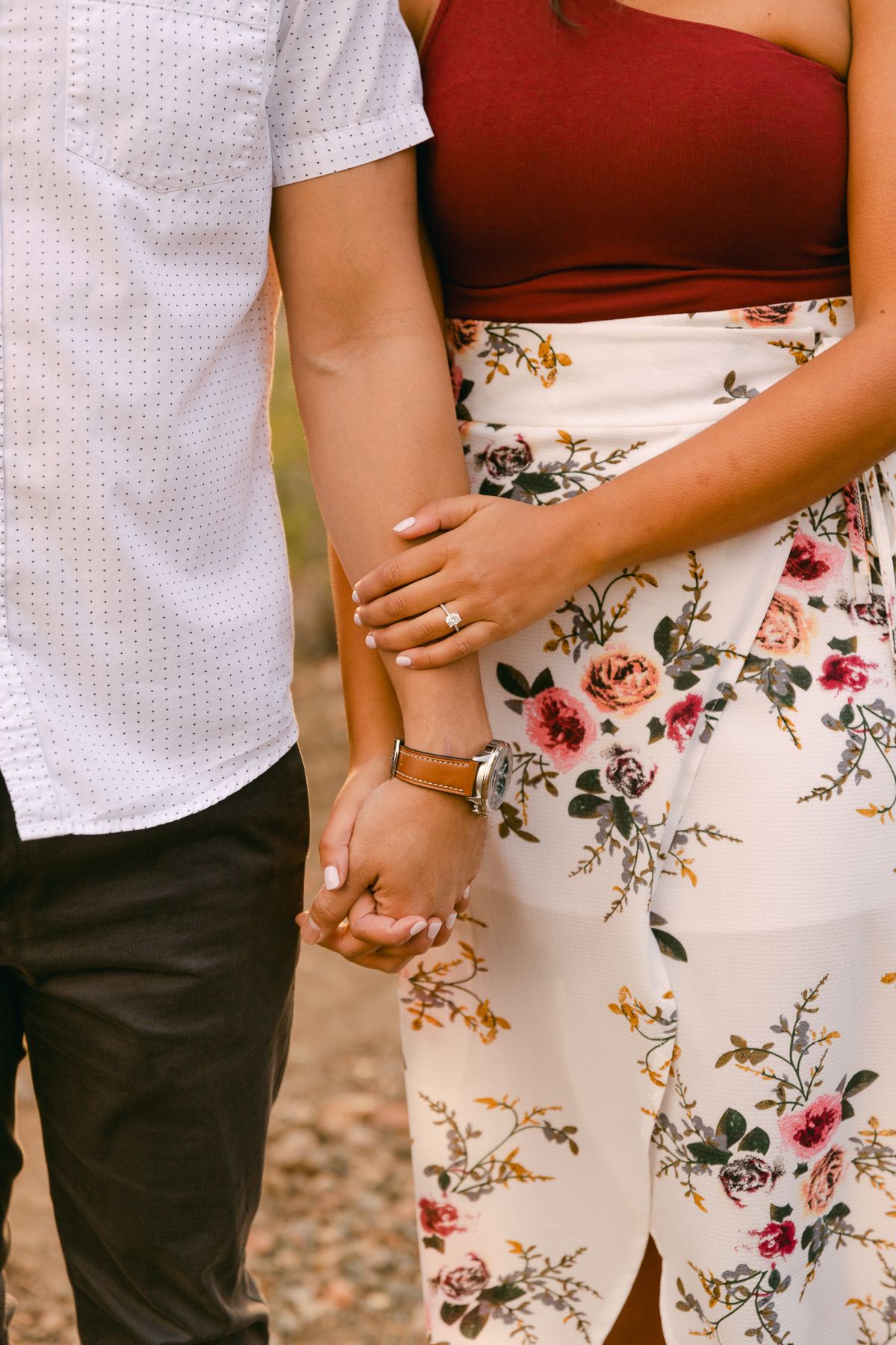 A couple is standing together and holding hands. One person is in a red top and white floral skirt and the other is in a white shirt.