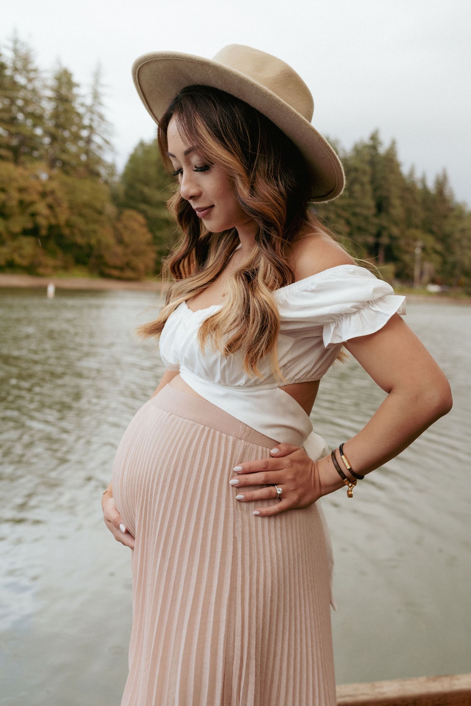 Expecting mother in hat cradling belly by water, rustic PNW lake setting in Oregon, warm natural tones.