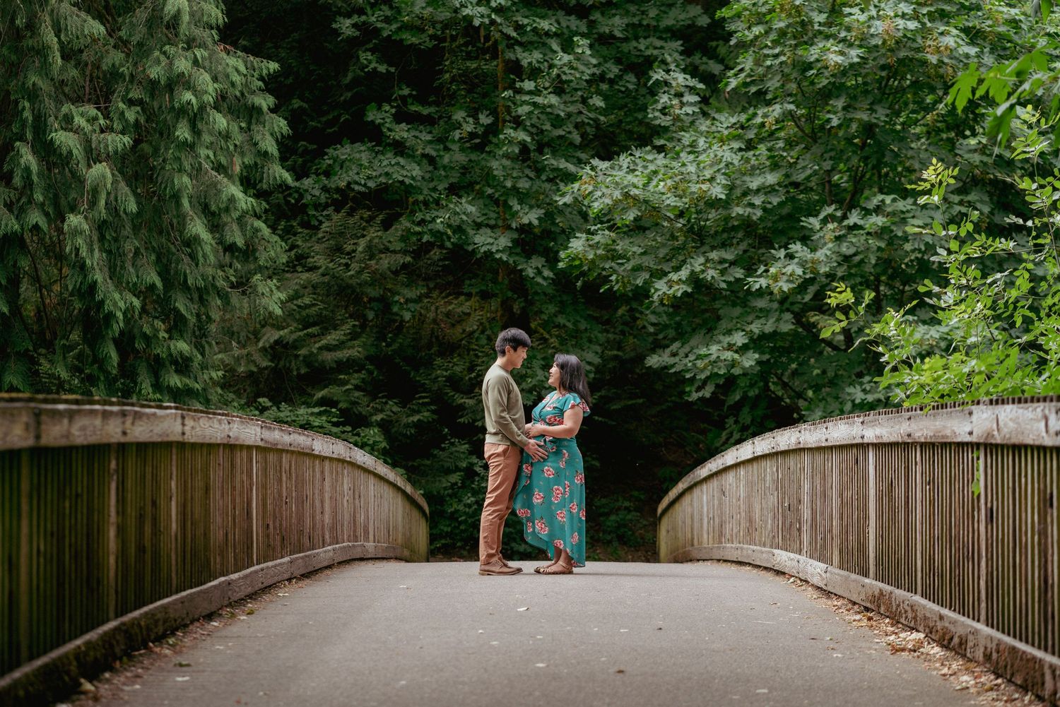Expecting parents holding hands on curved wooden bridge, Pacific Northwest park maternity photography.