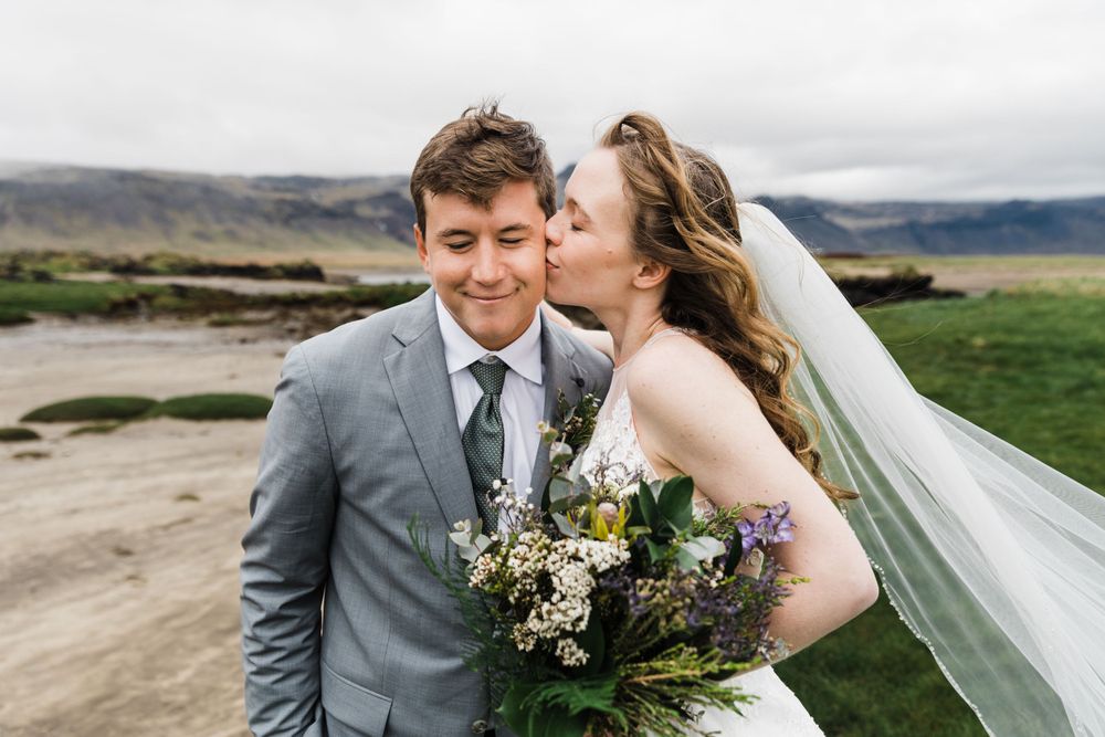 A joyful wedding couple embraces in front of a scenic rural landscape with mountains and fields.