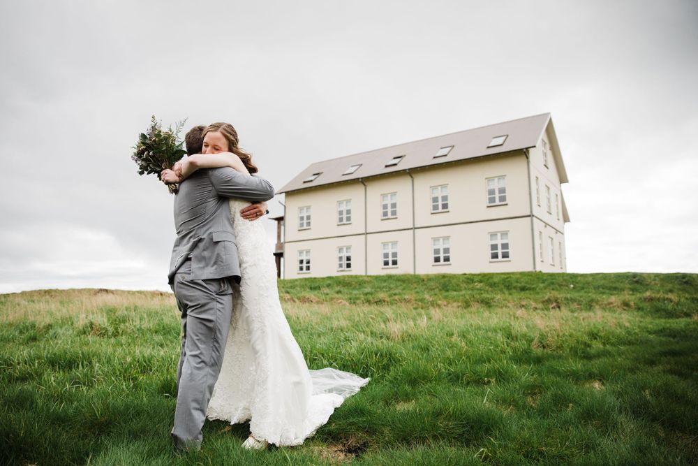 A couple embraces on grassy hillside with large building in background.