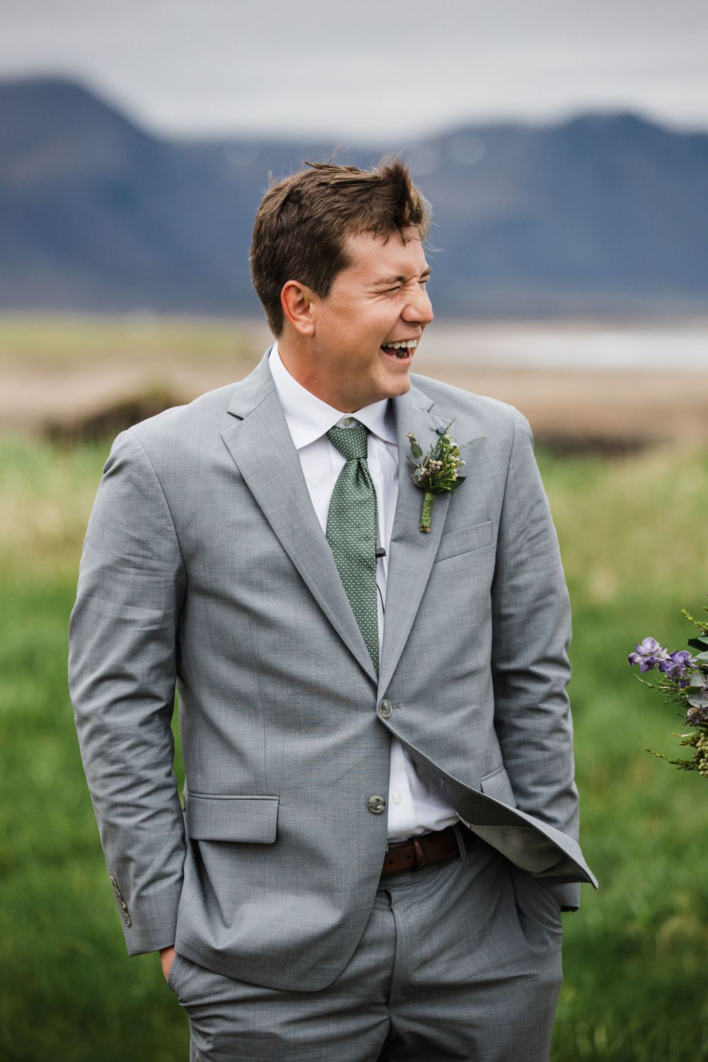 Person in grey suit with green tie laughing against mountainous backdrop.