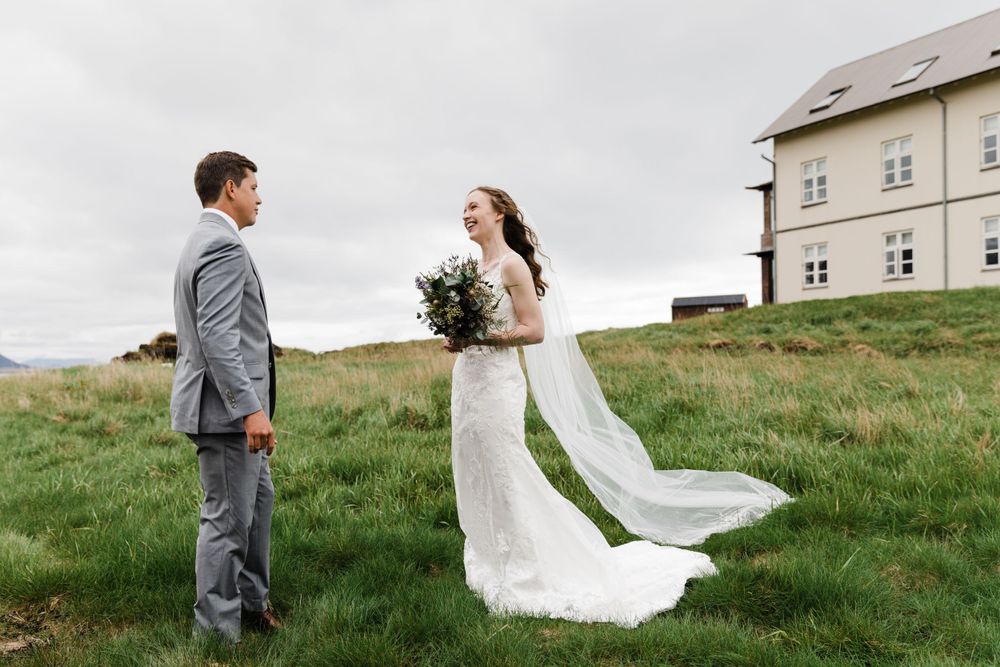 A couple stands in a grassy field with a light grey building in the background.