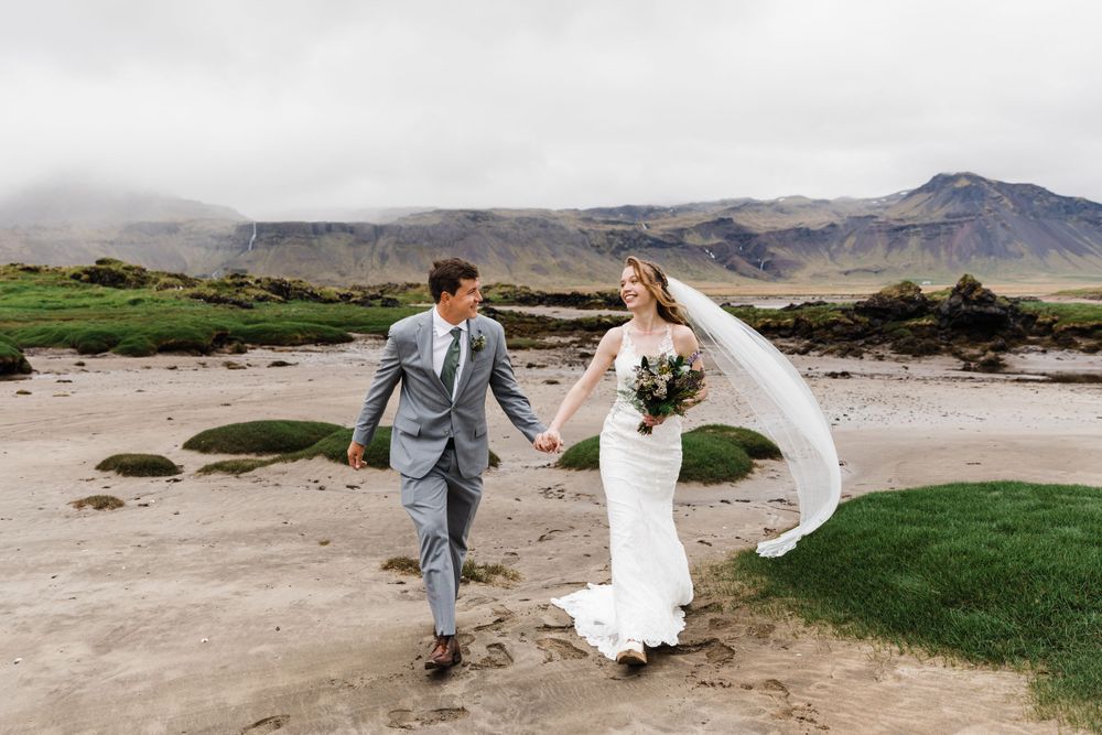 A couple walks hand in hand across a dramatic Icelandic landscape with mountains in the background.