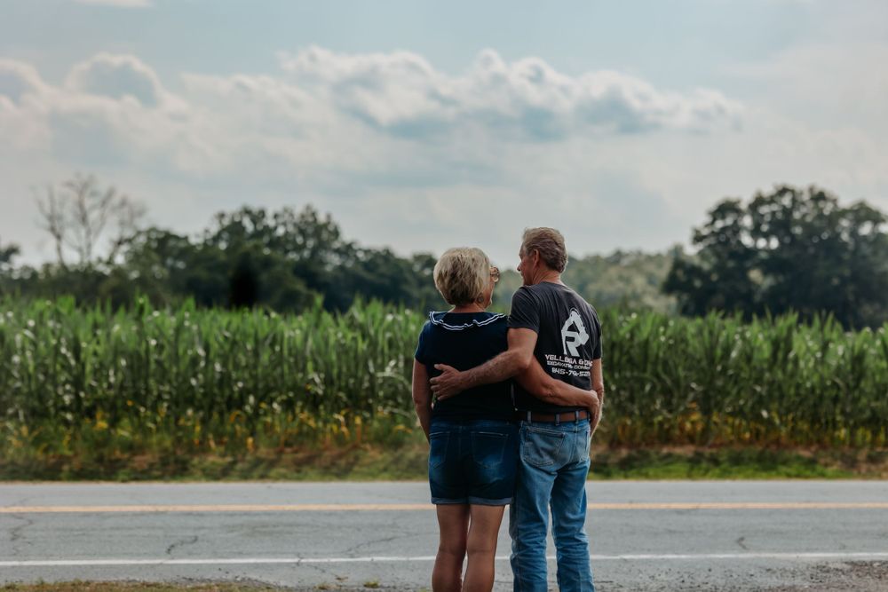 Two people embrace while looking at cornfield and cloudy sky.