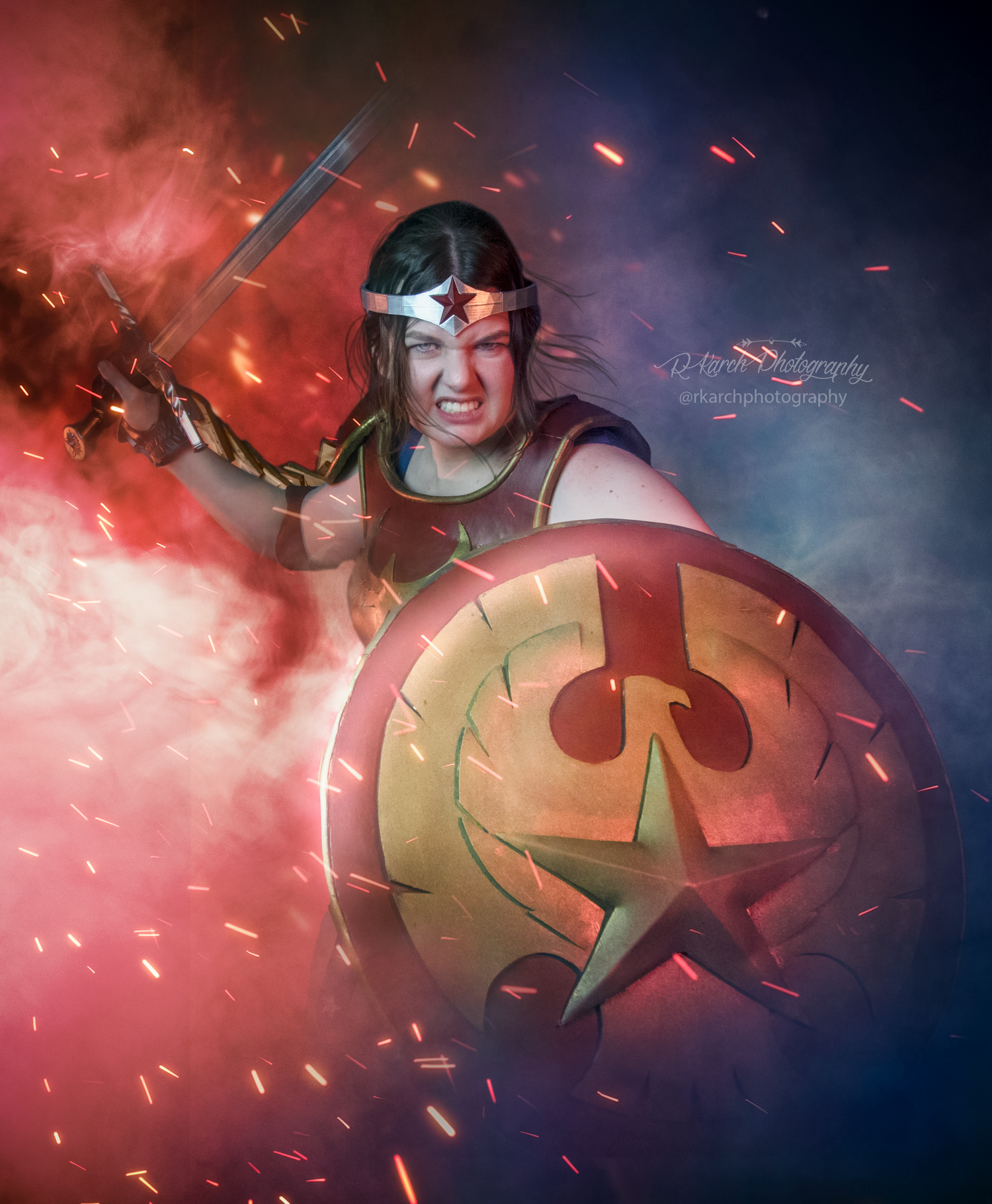 Warrior cosplay with decorative shield and headband posing dramatically in red and blue smoke.