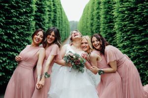Bridal party in pink dresses poses joyfully between rows of tall green hedges.