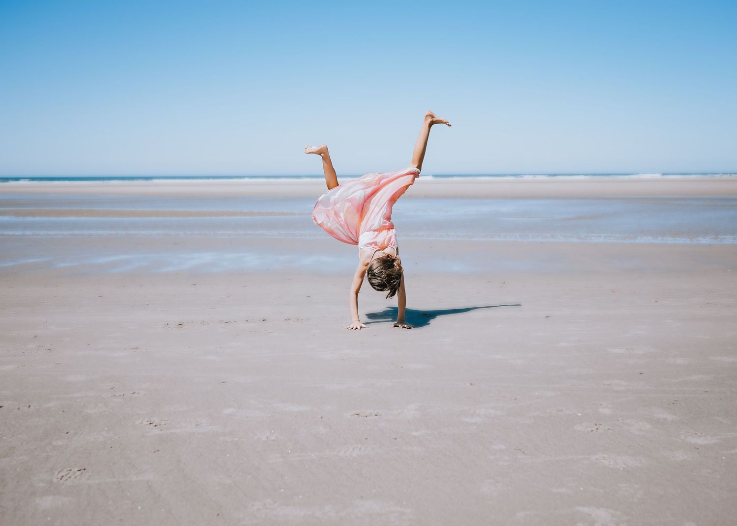 Person in pink dress does a cartwheel on wet sandy beach under blue sky.