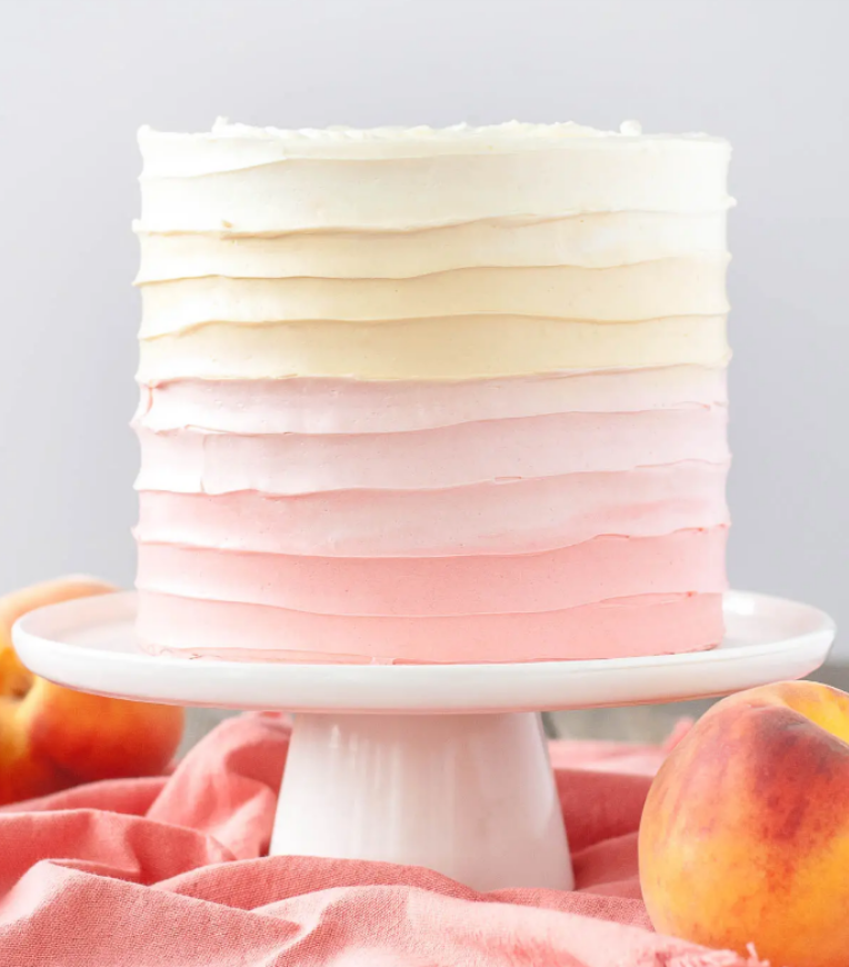 Ombre cake decorated in white to pink ruffled frosting on white cake stand with peaches.